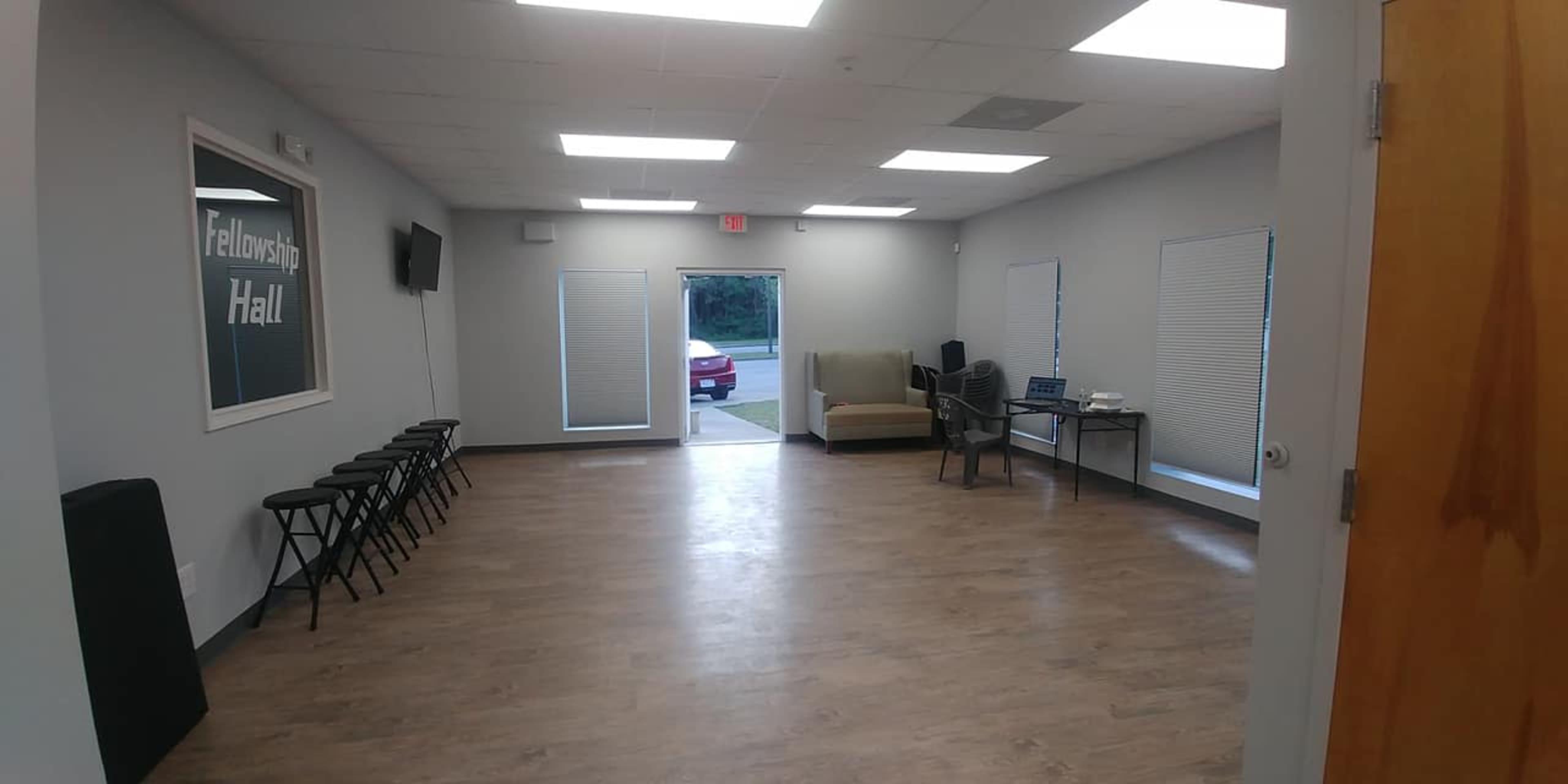 A spacious room with a few folding chairs lined up against one wall and a sofa, desk, and computer setup on the opposite side, with a door leading outside.