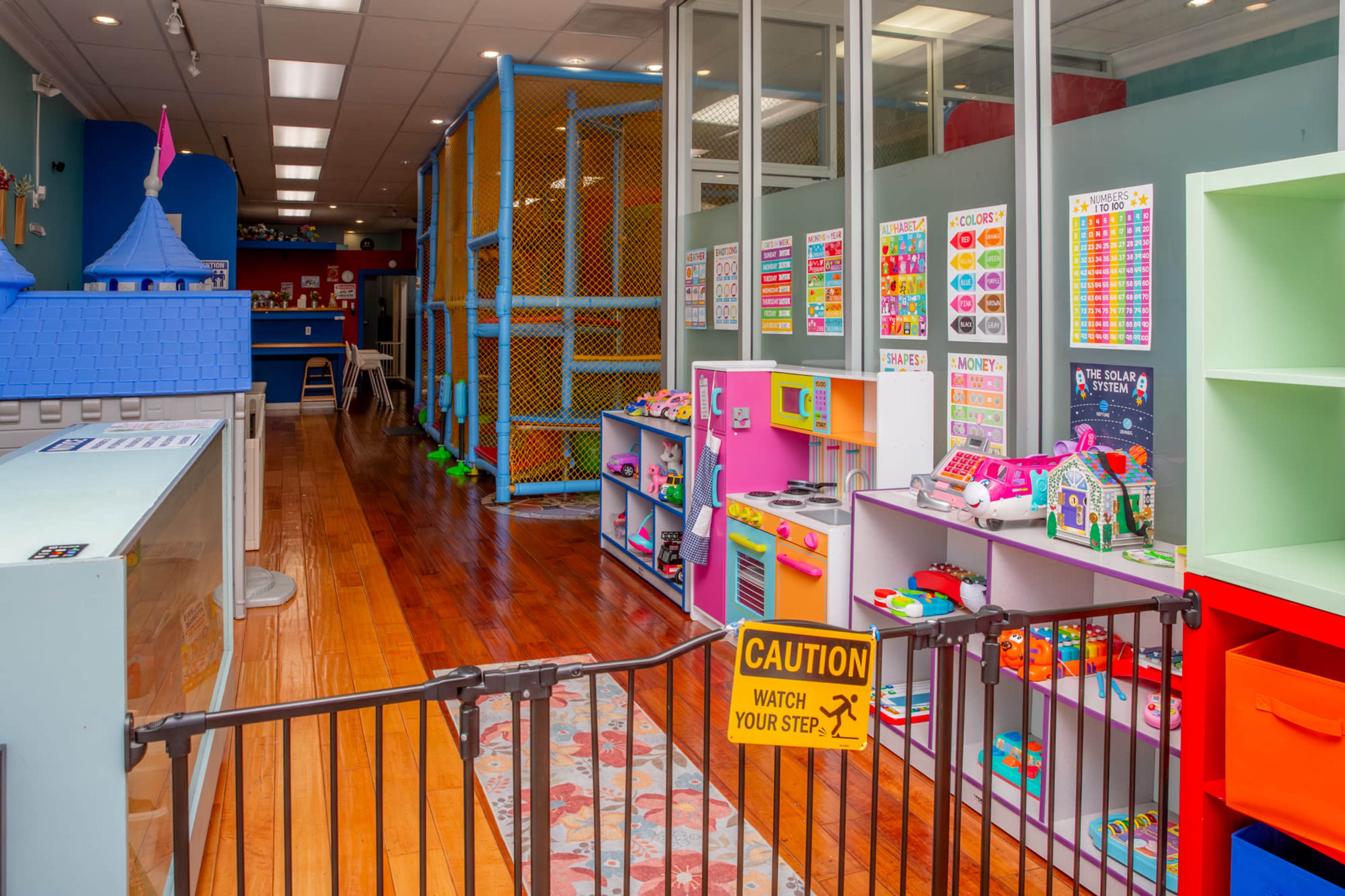 The image shows a colorful children's playroom with various toys, play structures, and safety signage.
