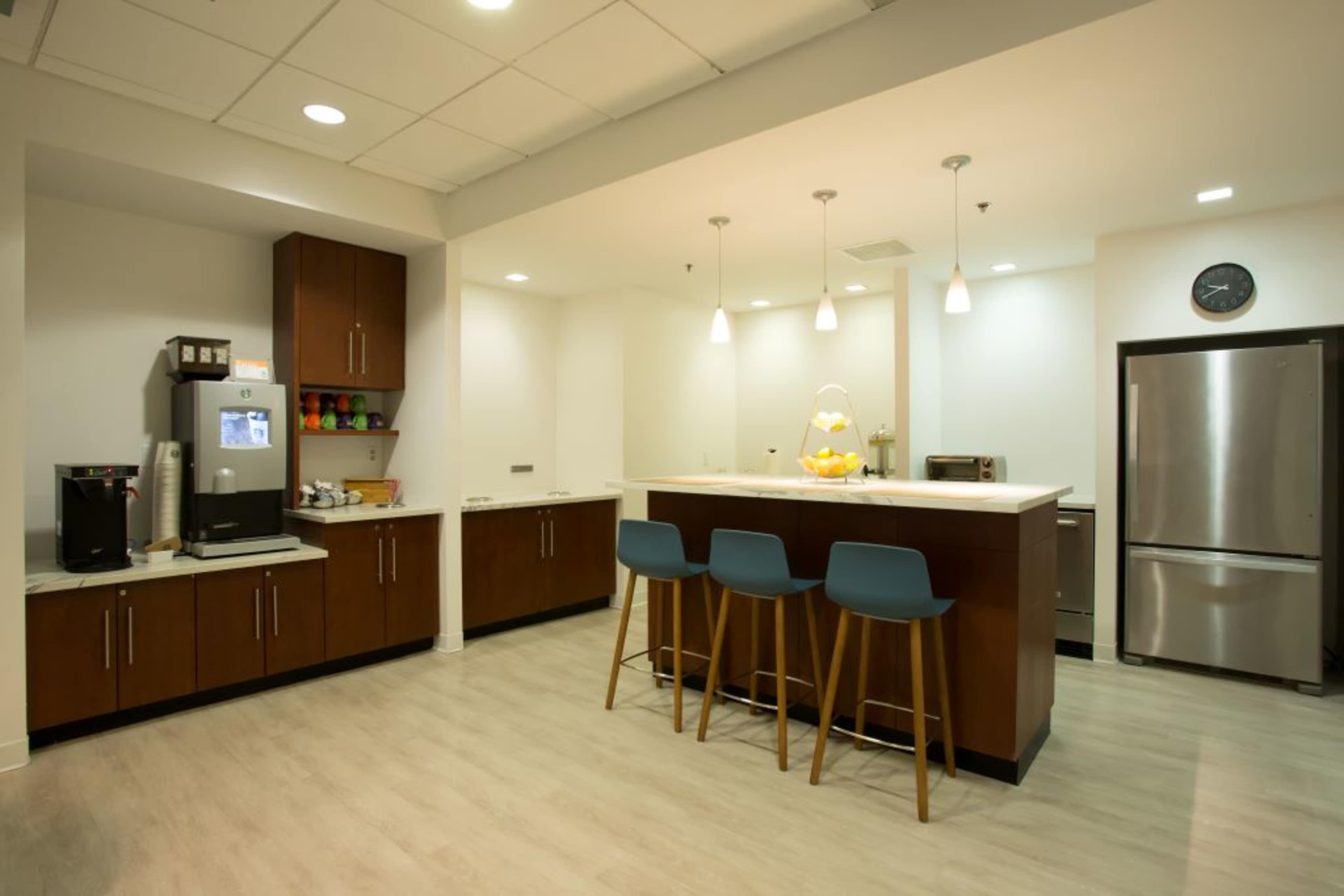 The image shows a modern kitchen with a bar counter, three blue stools, wooden cabinets, and stainless steel appliances.