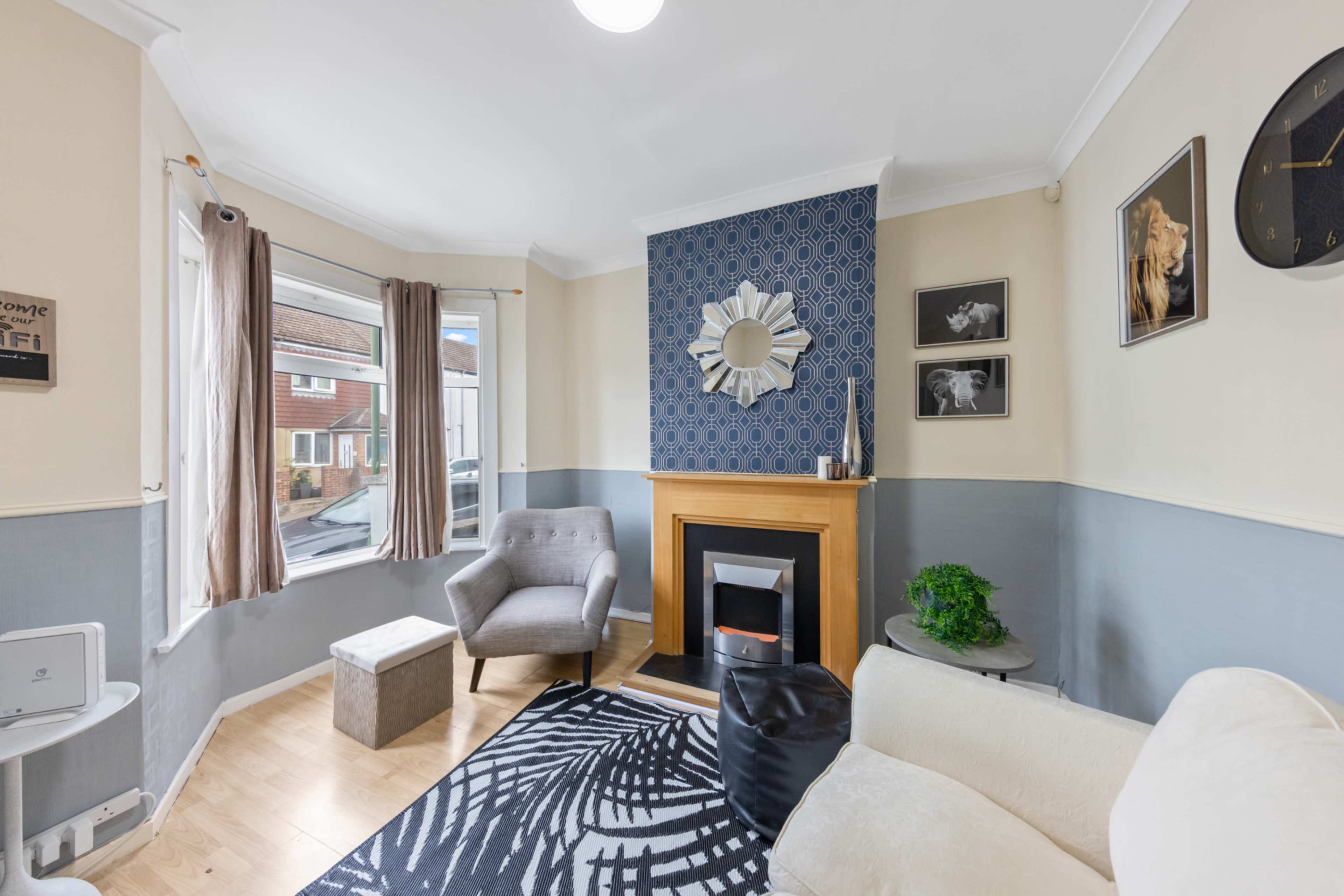 The image shows a small living room featuring a fireplace, a large mirror, a gray armchair, and a decorative rug on the wooden floor.