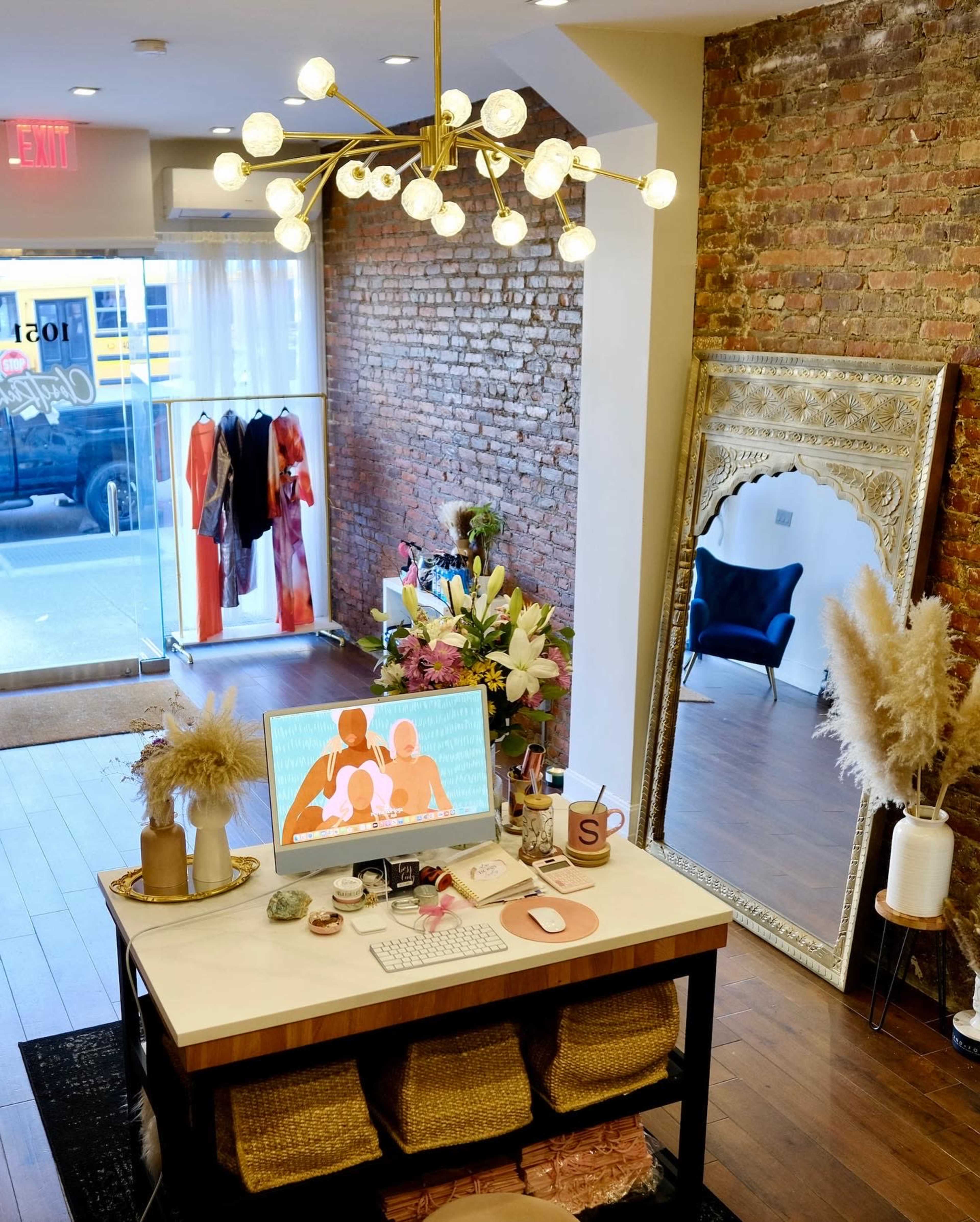 A warmly lit boutique with a brick wall, a display table featuring a computer and decorative items, and a large mirror, alongside clothing and floral arrangements.