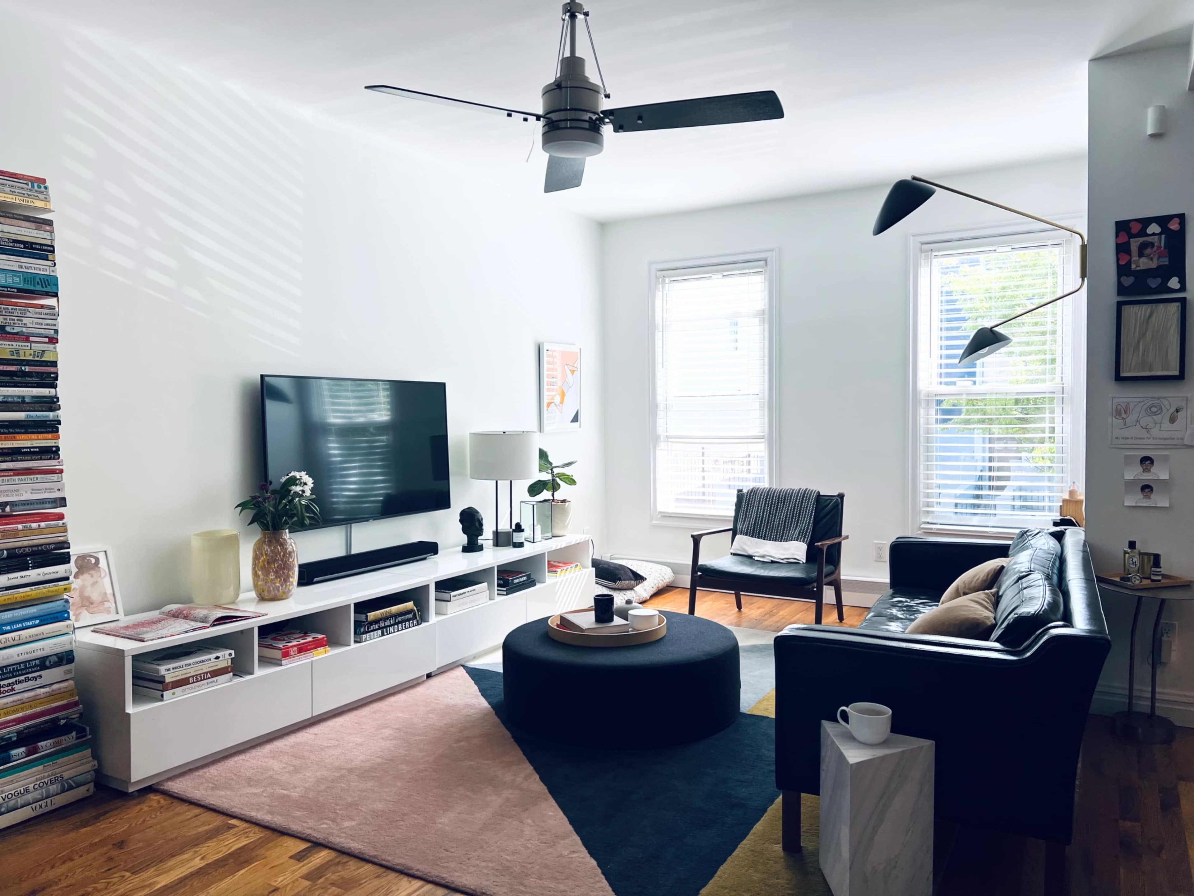 A modern living room featuring a television mounted on the wall, a black leather sofa, a round coffee table, and a bookshelf stacked with books.
