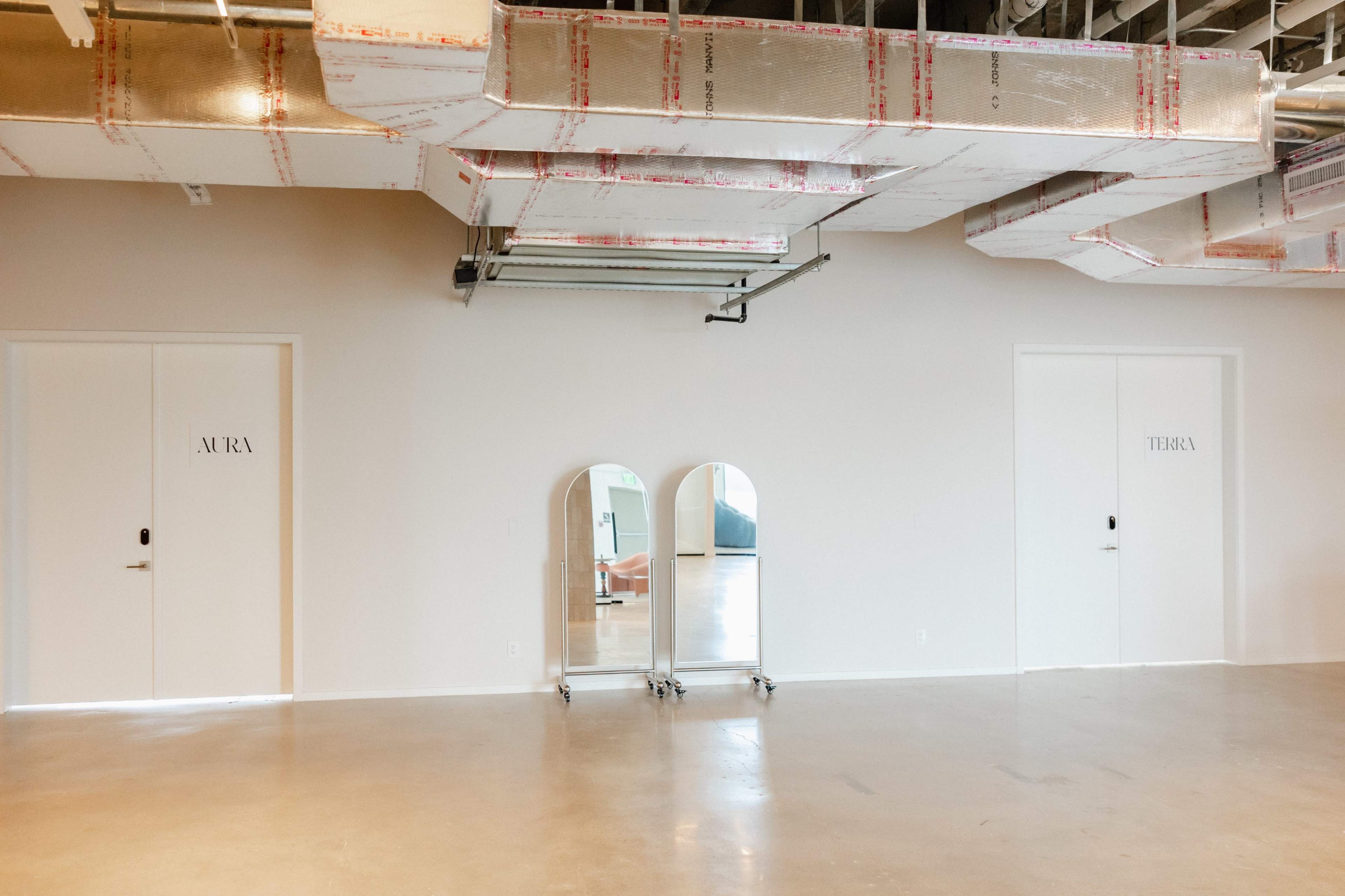 The image shows a minimalist interior space featuring three doors labeled "AURA" and "TERRA," with a mirror on wheels positioned in between.