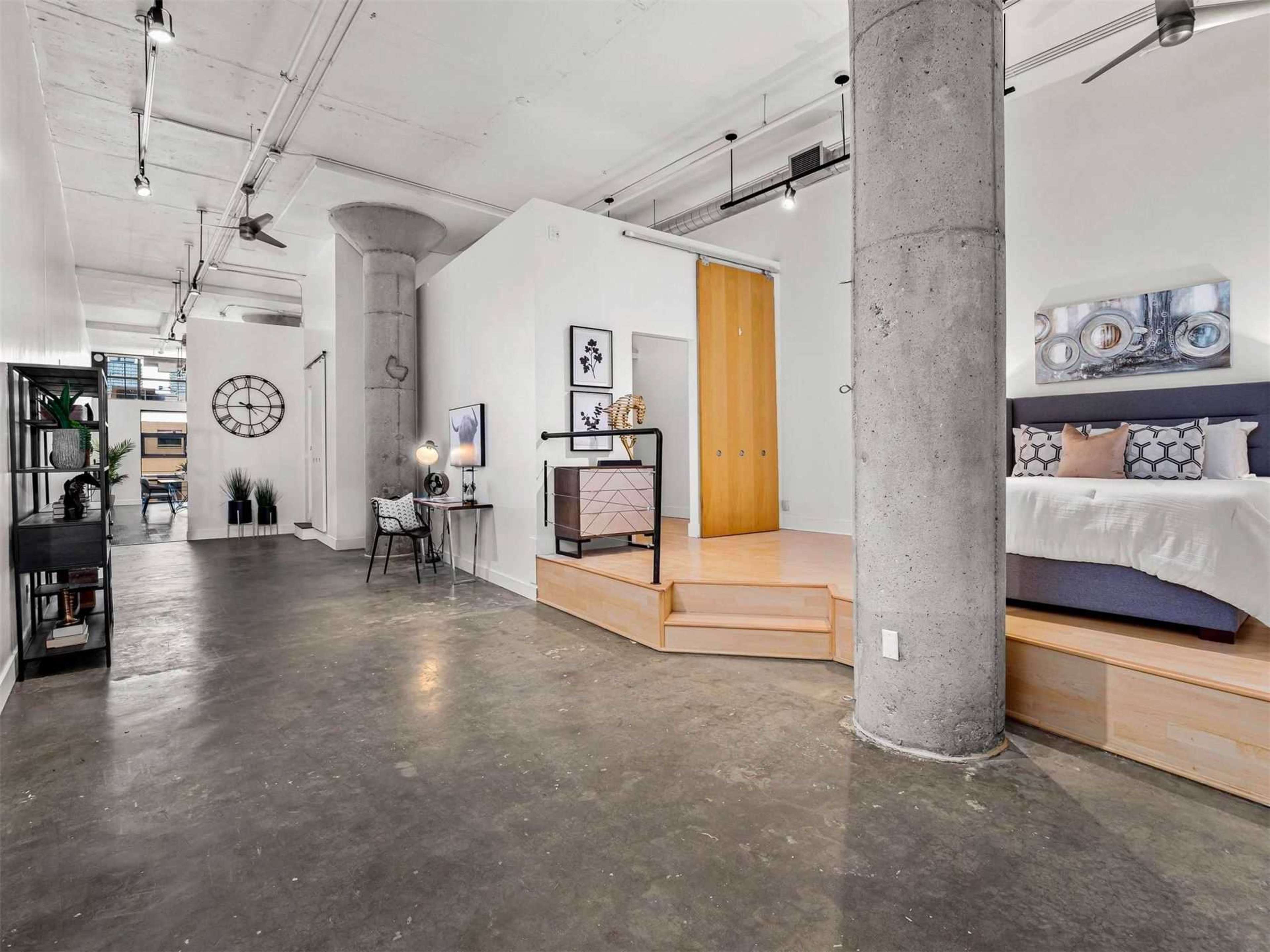 The image shows a spacious, modern interior featuring a raised platform with a bed, a seating area, and exposed concrete columns.