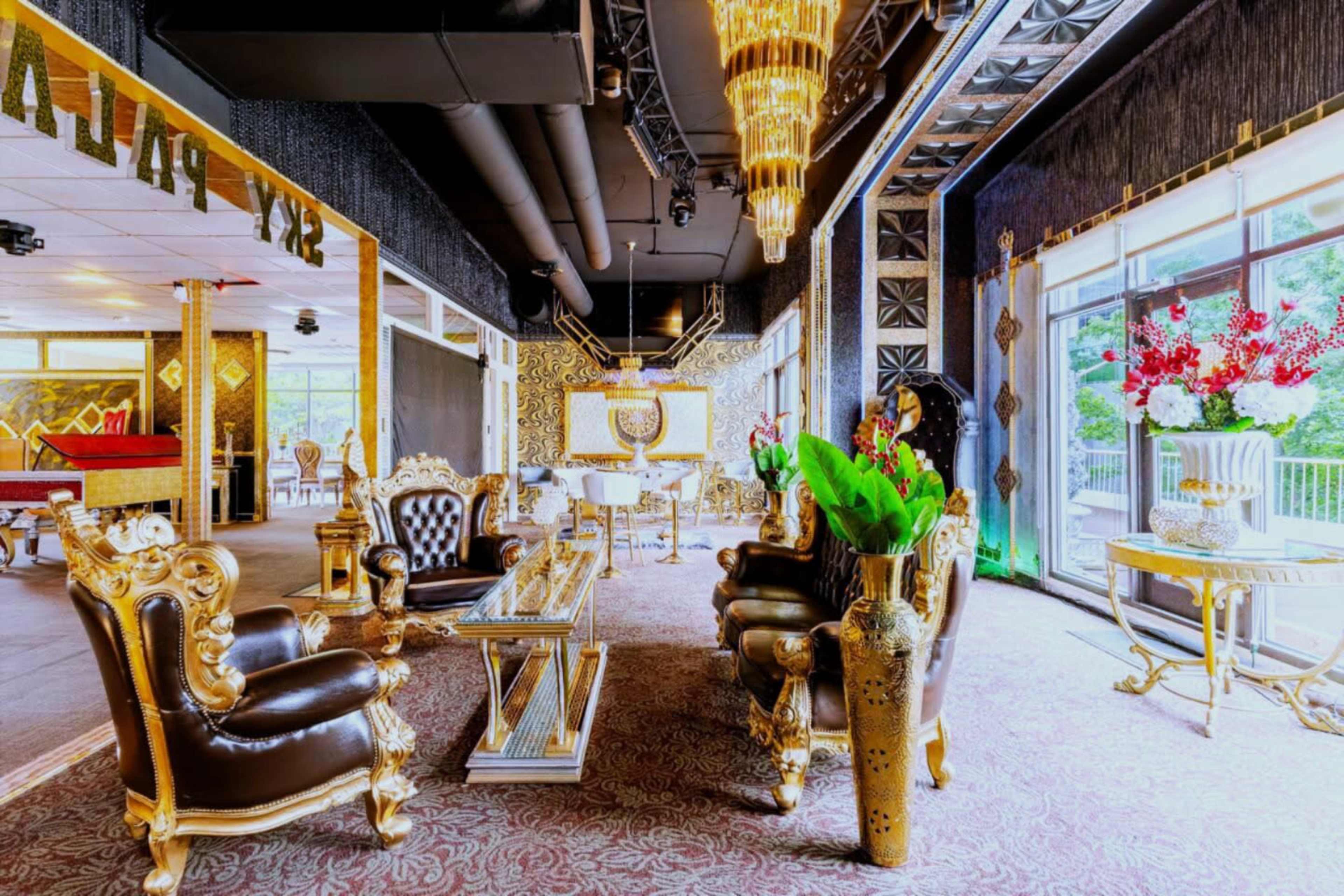 The image shows an opulent interior space featuring gold-accented furniture, including ornate chairs and vases, and large windows adorned with decorative plants and floral arrangements.