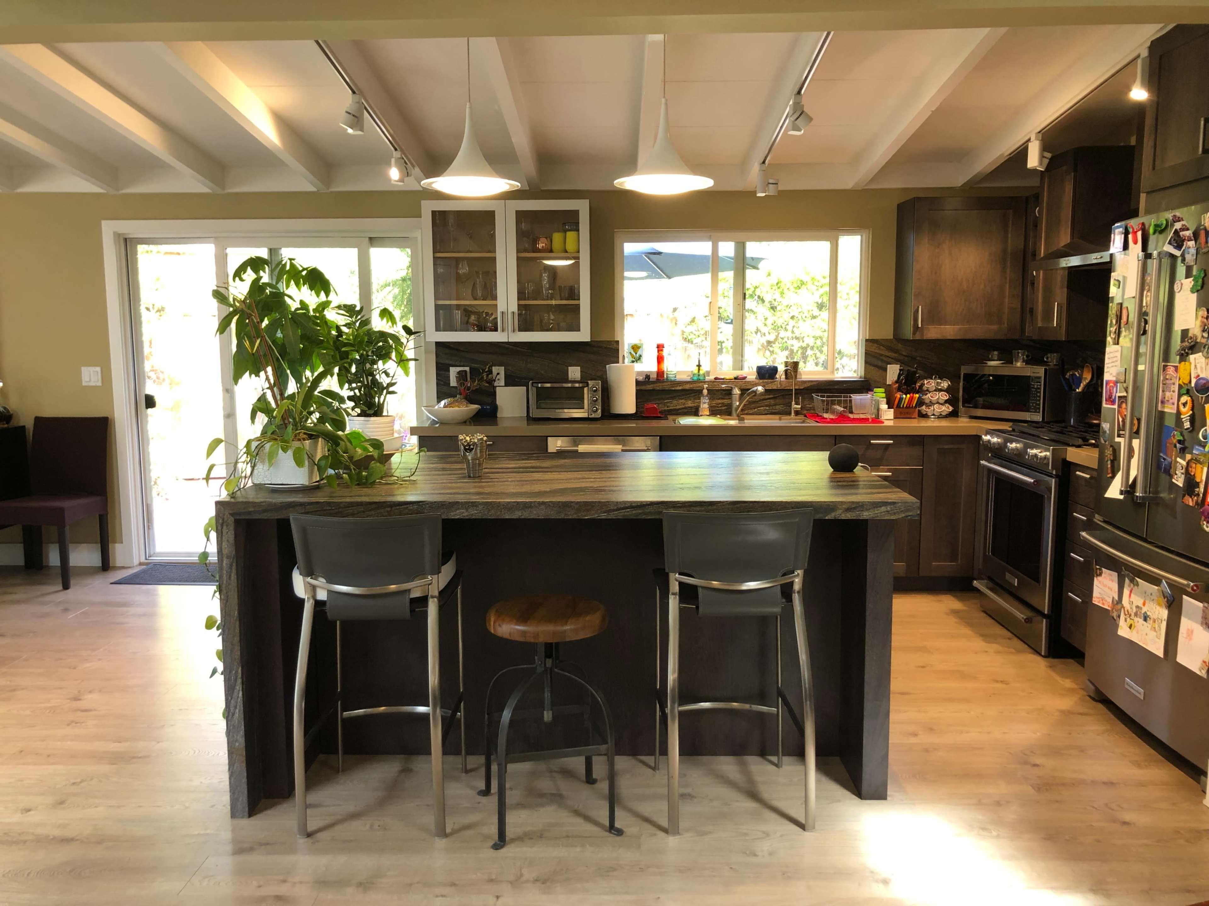 The image shows a modern kitchen with a central island, featuring bar stools, wooden cabinets, and large windows that allow natural light to enter.