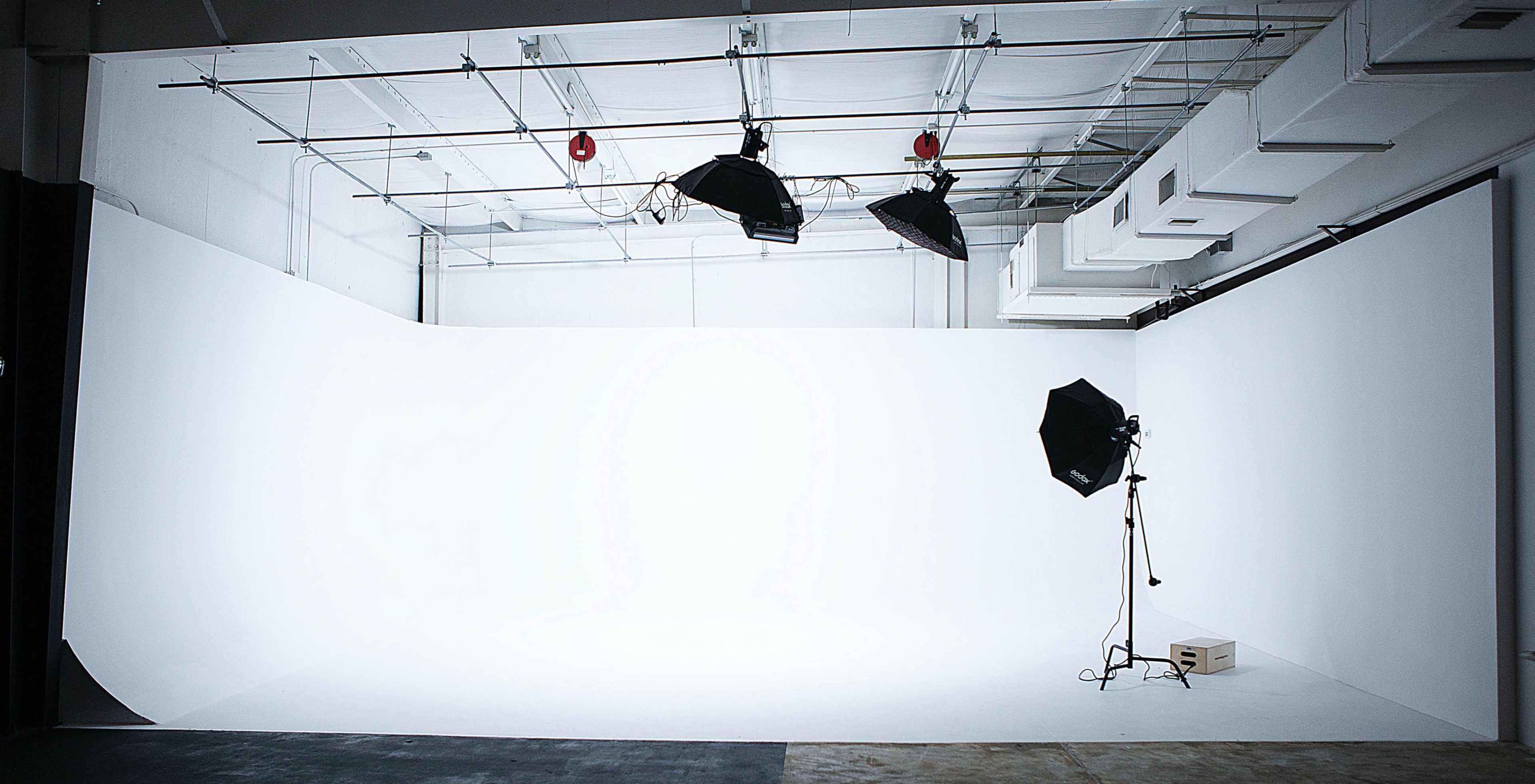 A photography studio with a large white backdrop, two overhead lights, and a softbox on a stand.