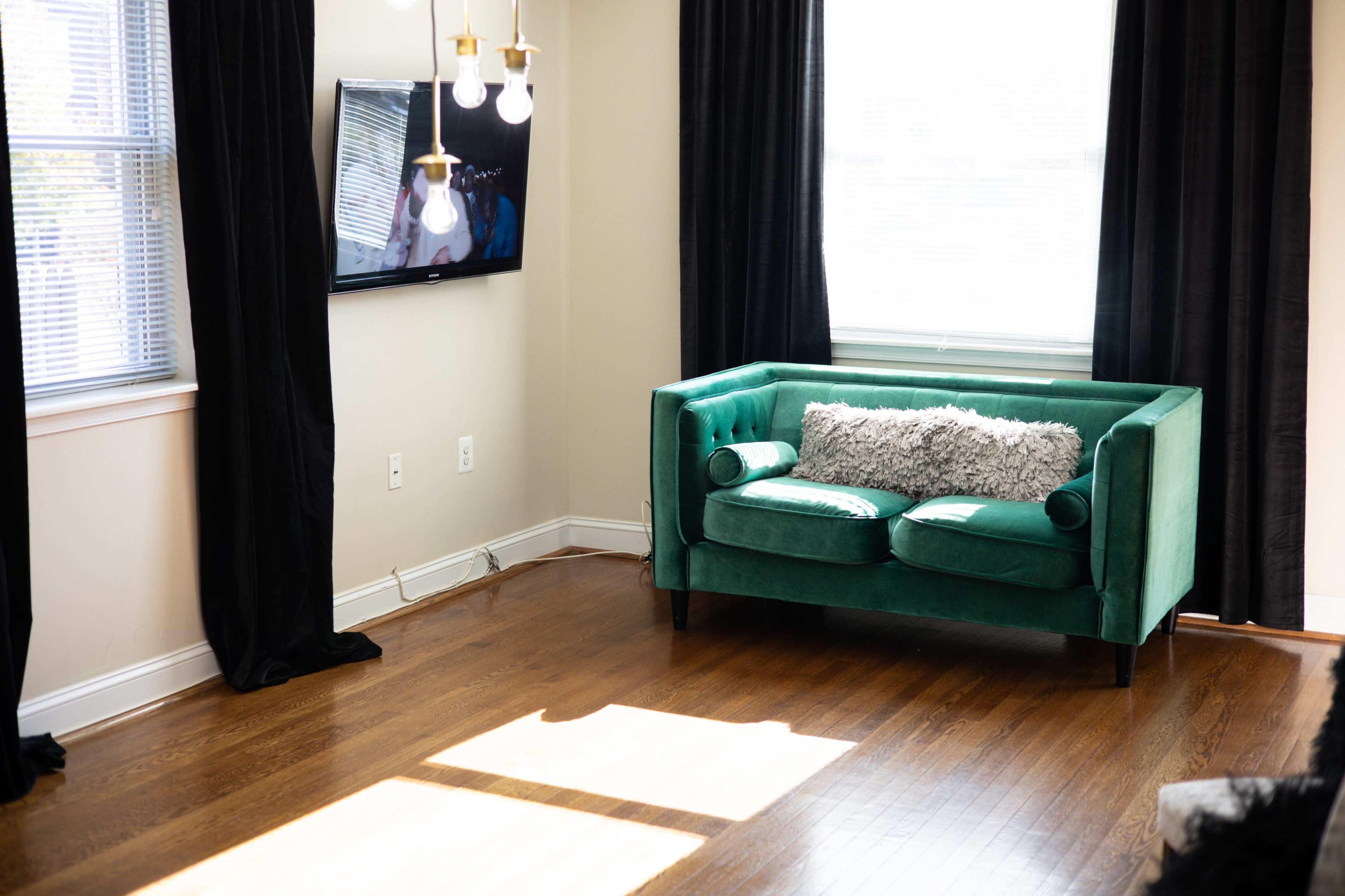 A green couch with decorative pillows is positioned in a sunlit corner of a room with hardwood floors and large windows.