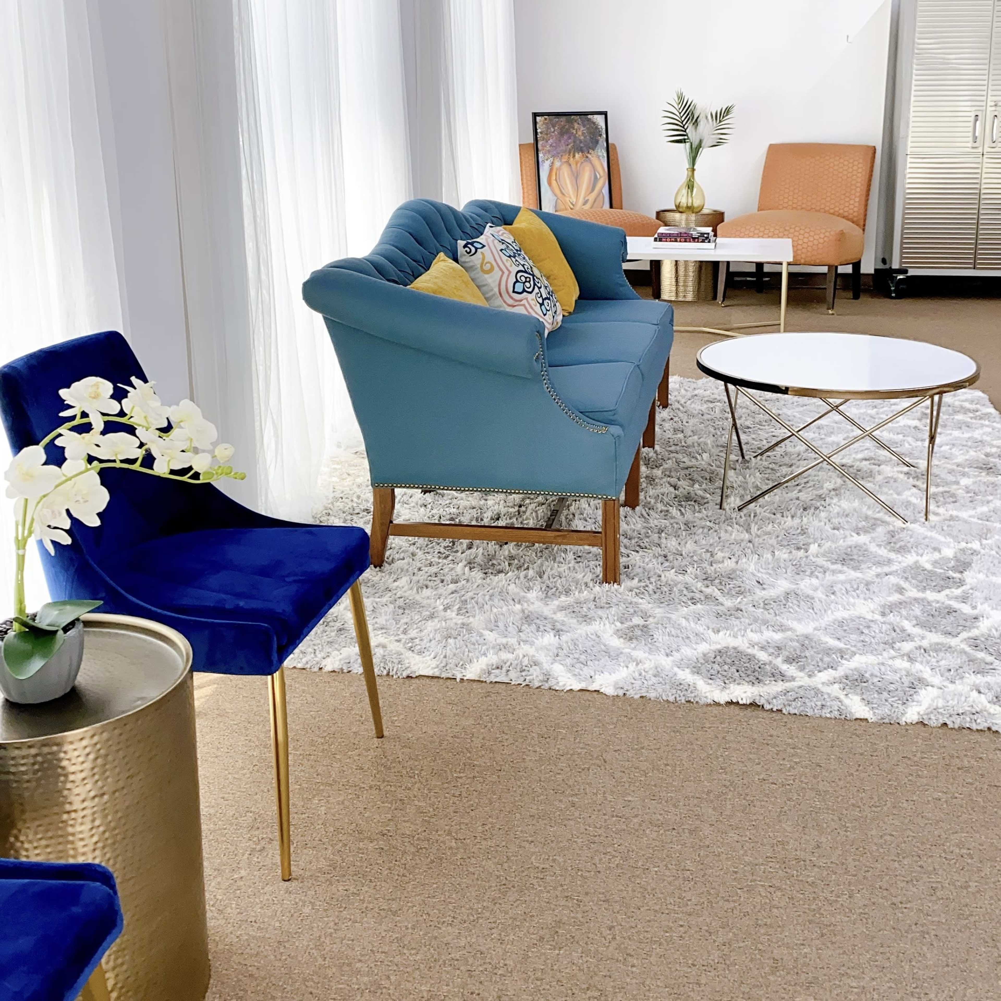 A living room with a blue upholstered sofa, a white circular coffee table on a textured rug, and two chairs alongside a gold side table with a potted plant.