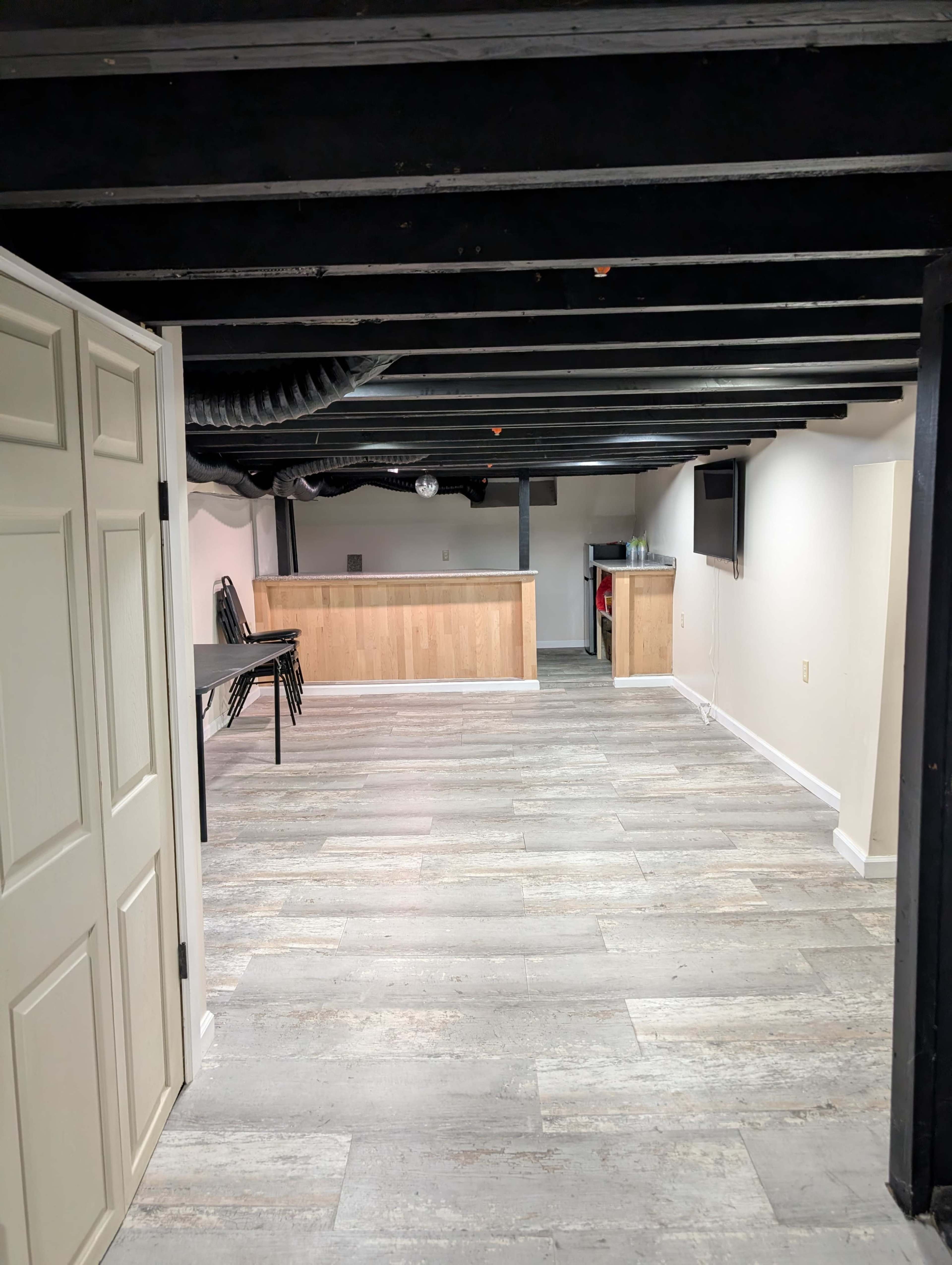 The image shows a spacious basement with wooden flooring, a bar area in the background, and a door on the left side.