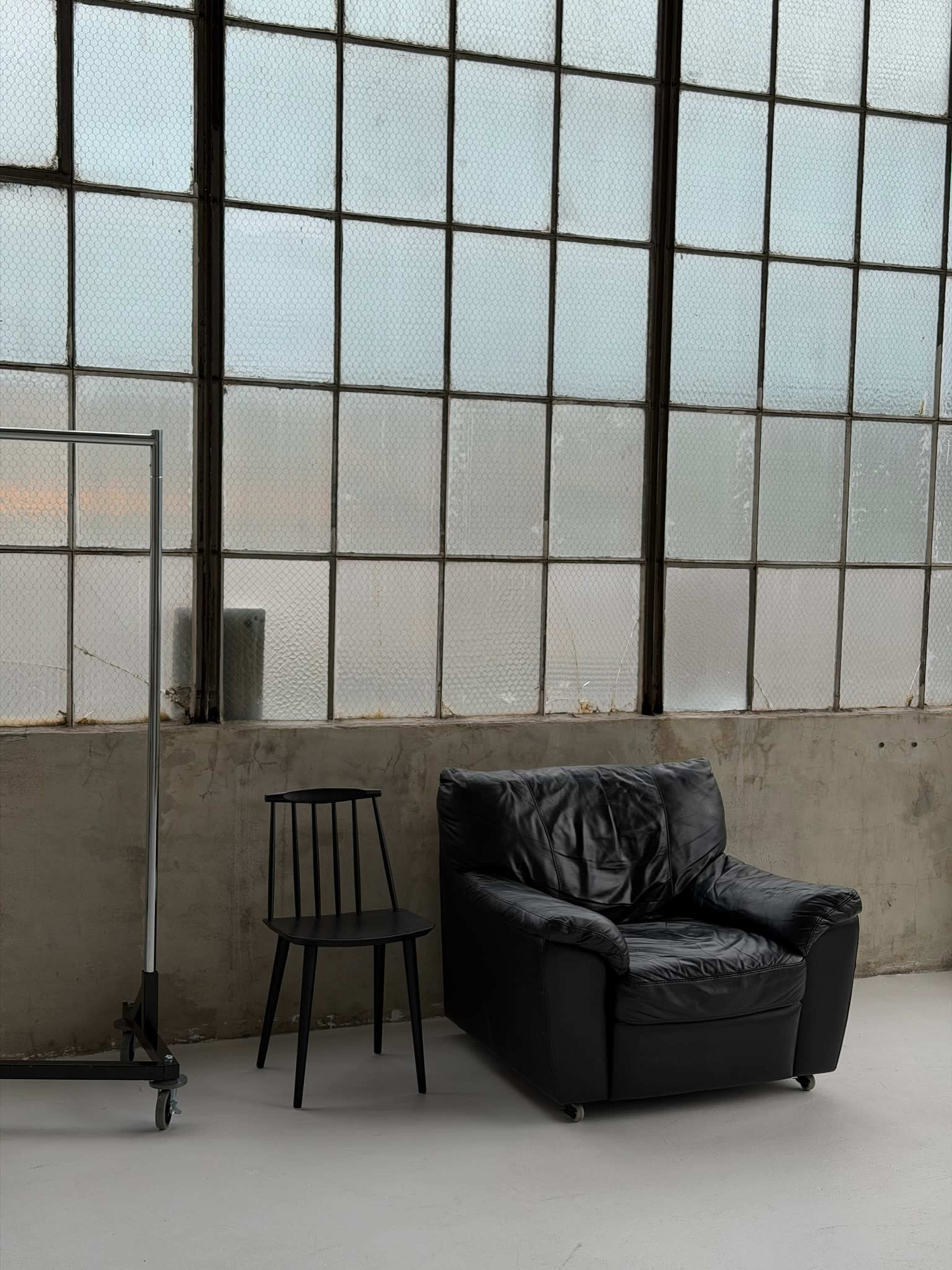 The image shows a black leather armchair next to a black wooden chair, set against a wall of large, paneled windows that diffuse natural light.