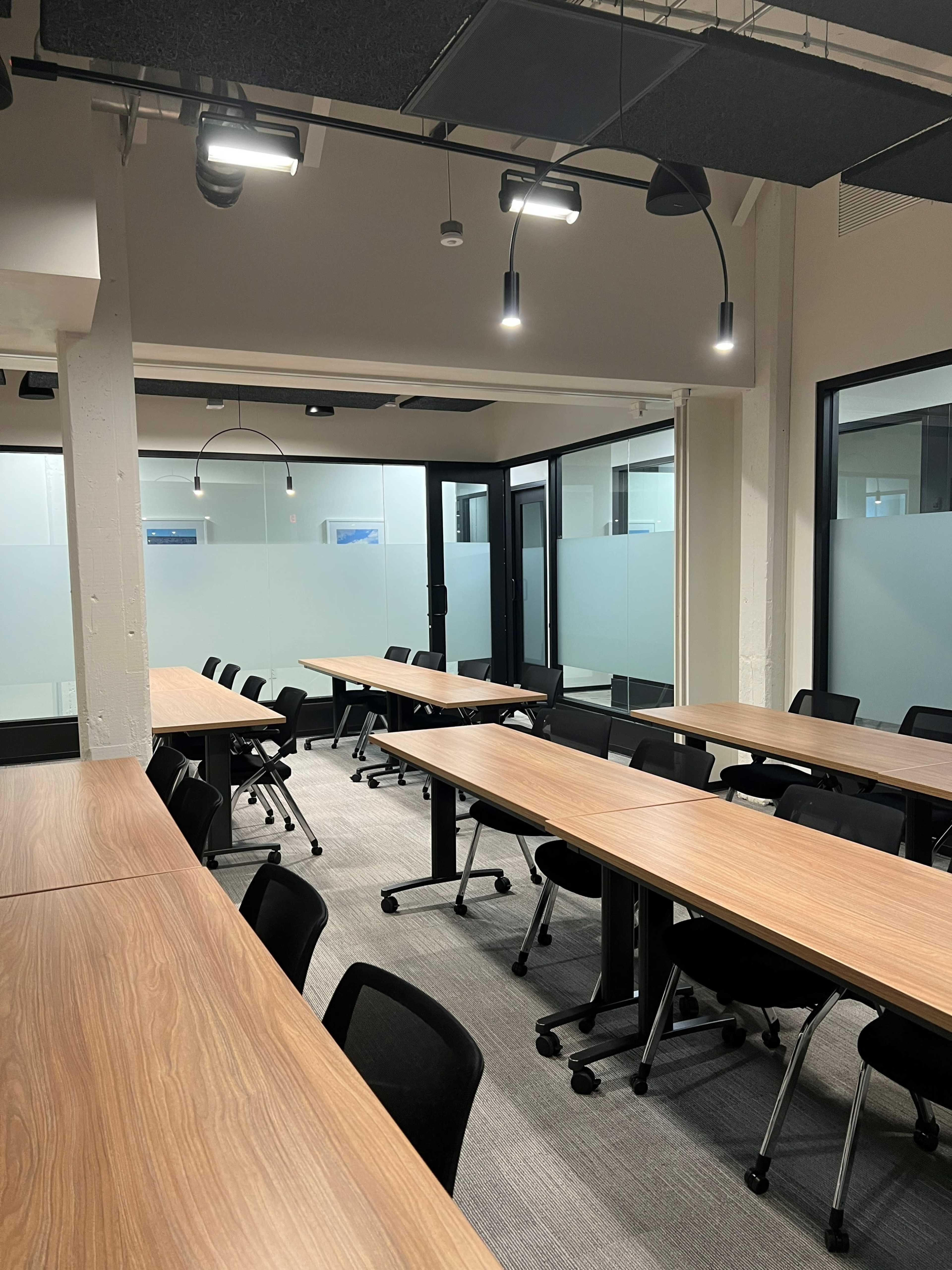 The image shows a modern conference room with several rows of long wooden tables and black rolling chairs, illuminated by overhead lights.