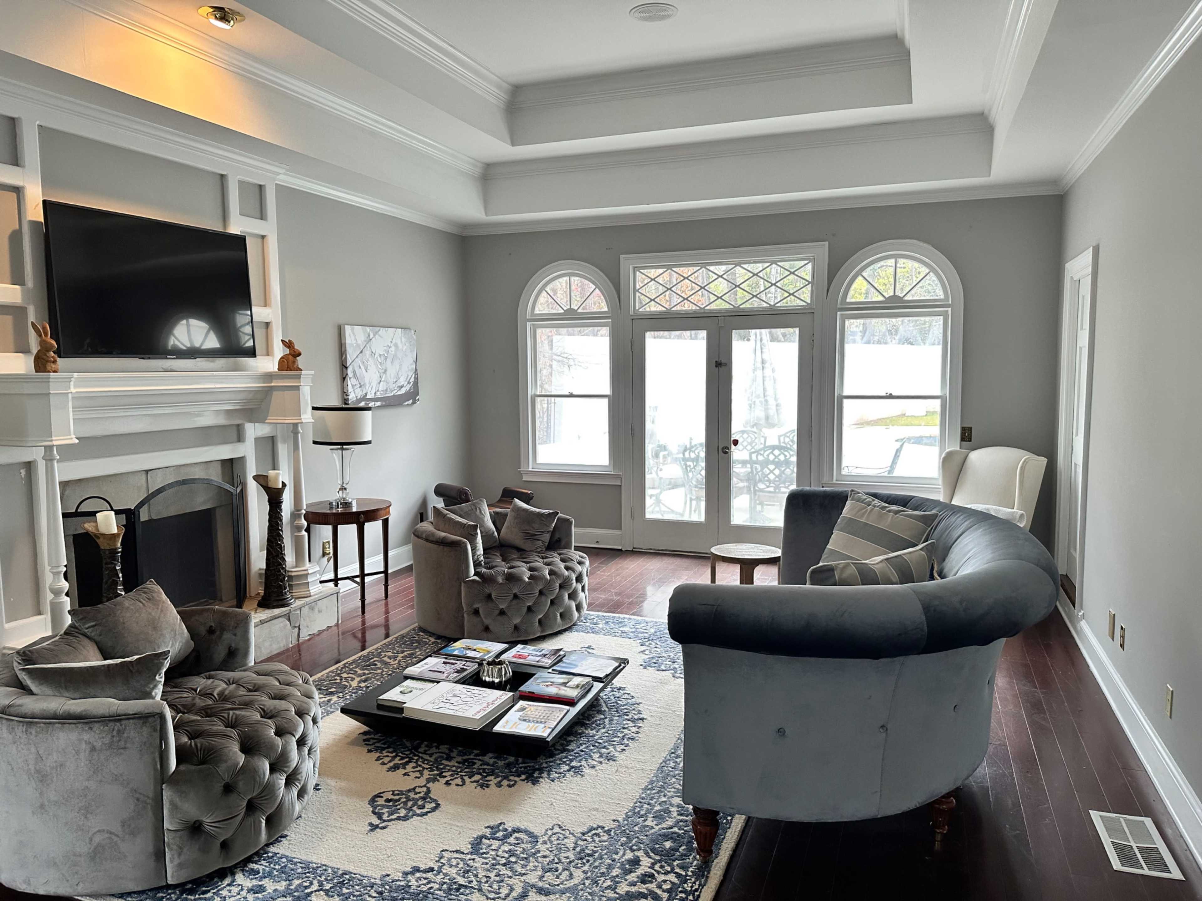 The image shows a modern living room featuring a circular seating arrangement with two tufted chairs, a fireplace, and large windows leading to a patio.