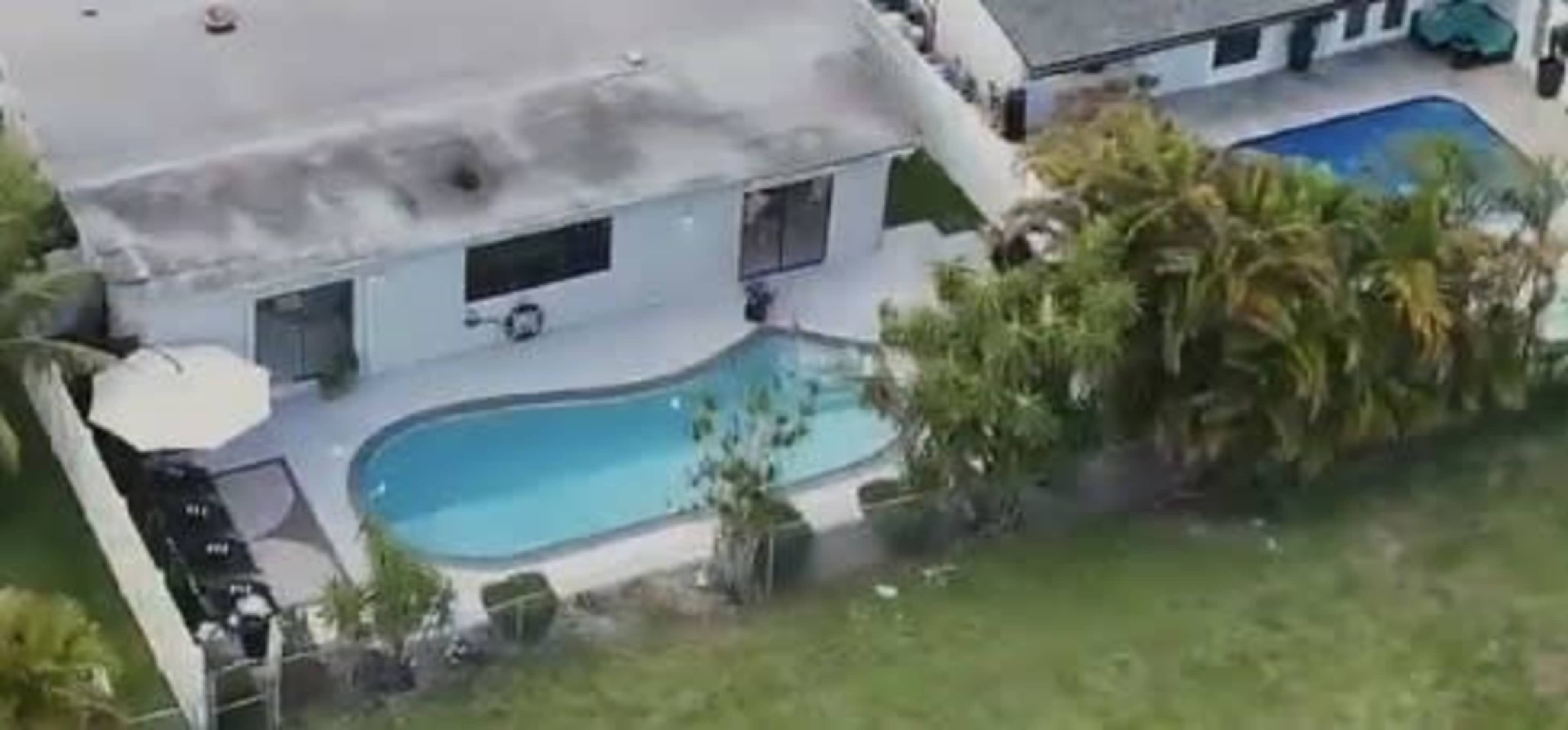 The image shows a house with a curved swimming pool surrounded by a grassy area and trees, with another pool visible at an adjacent property.