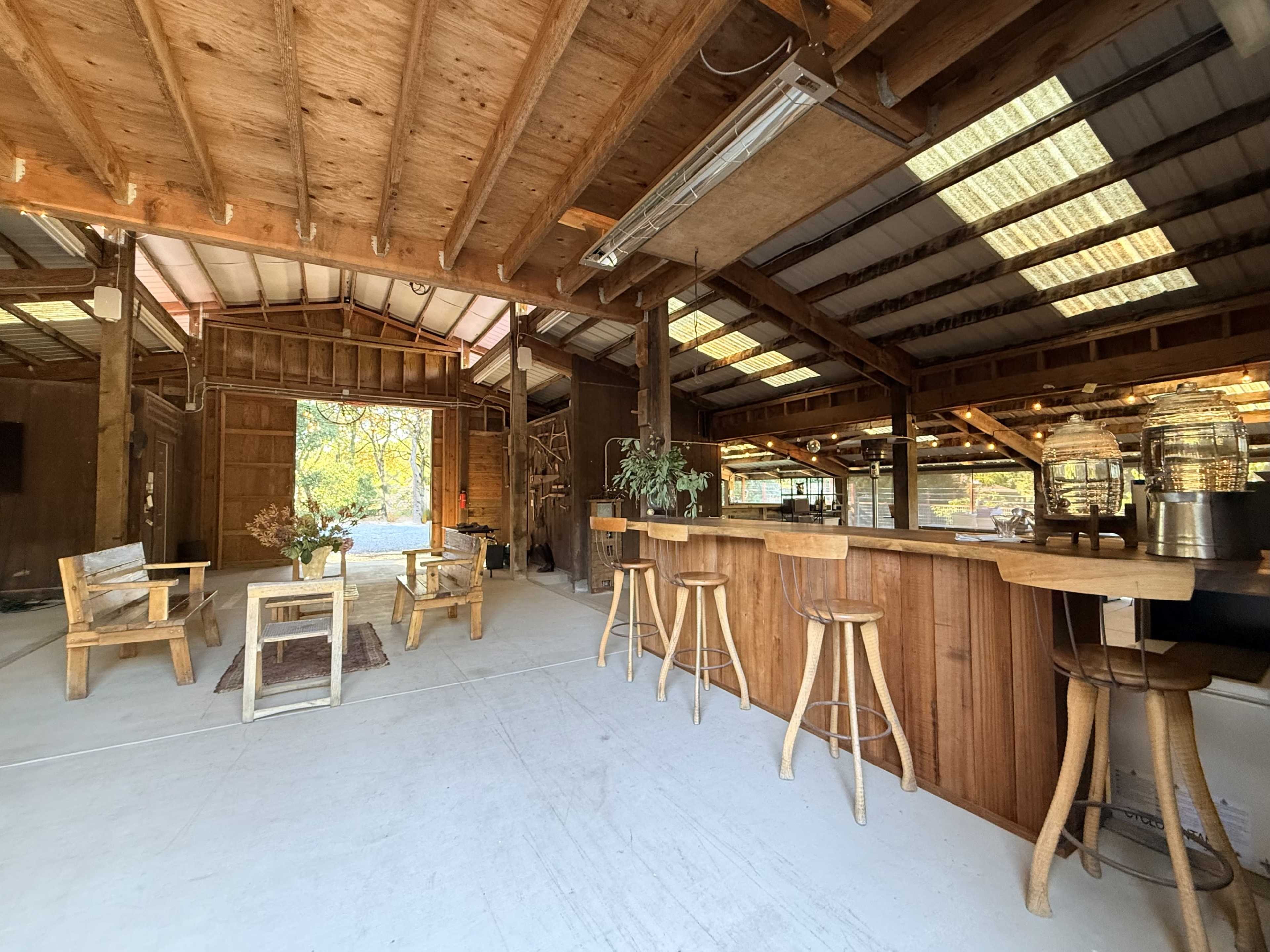 The image shows a spacious interior of a wooden structure with a bar counter, seating areas, and an open door leading to an outdoor view.