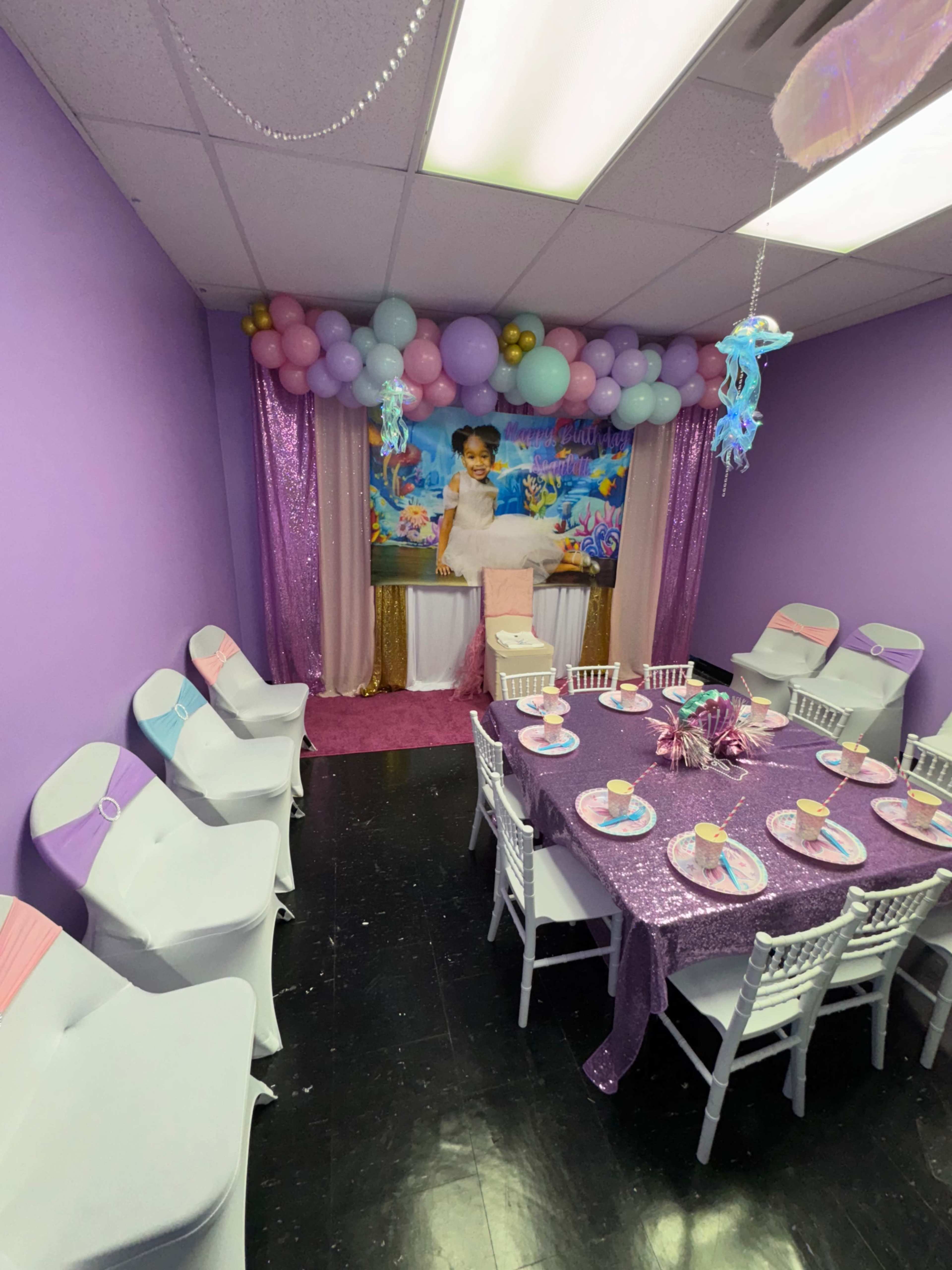 The image shows a decorated party room with a purple theme, featuring a table set with plates and decorations, surrounded by white chairs and balloons in pastel colors.