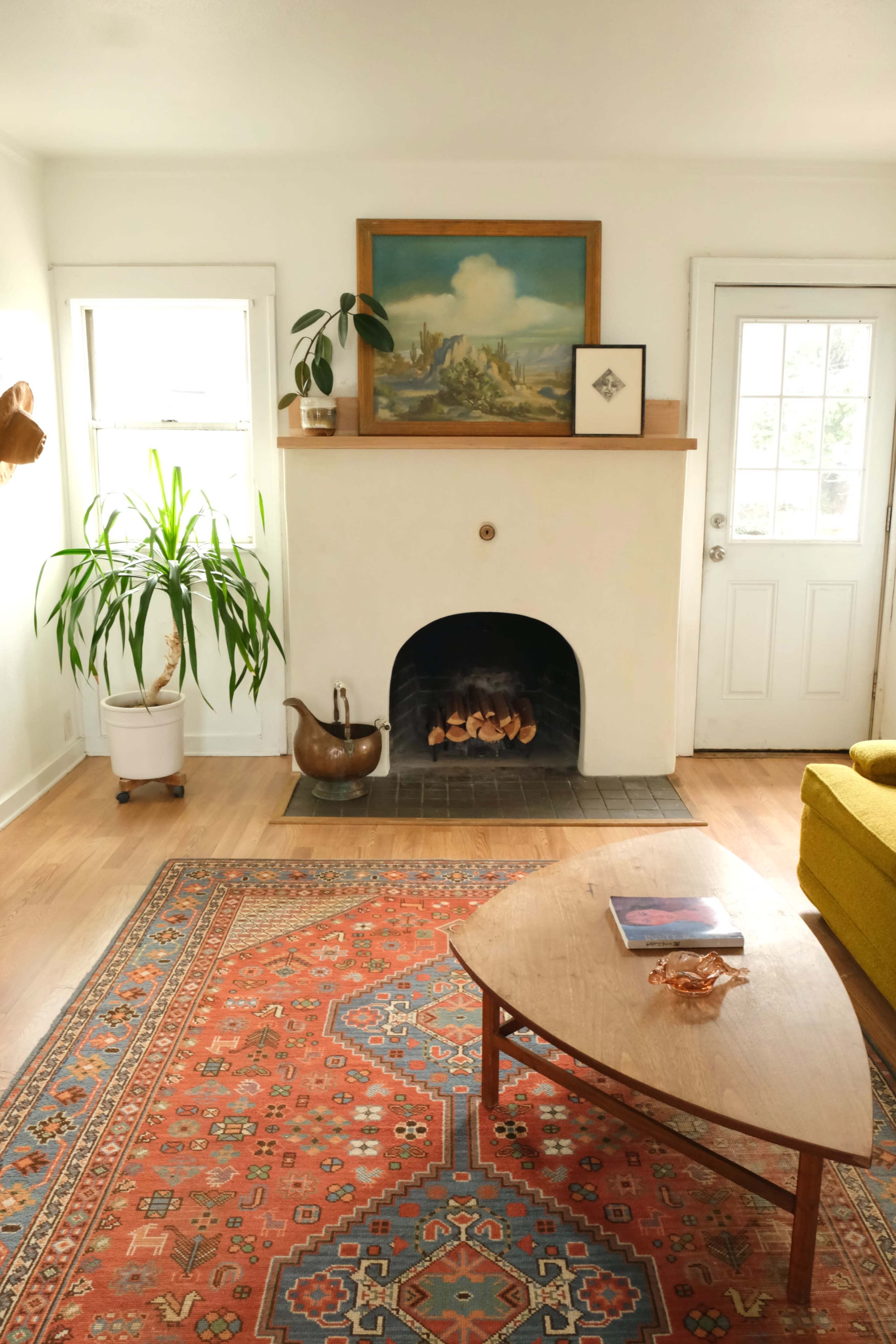 A cozy living room with a fireplace, a potted plant, an artwork on the wall, and a wooden coffee table on a patterned rug.