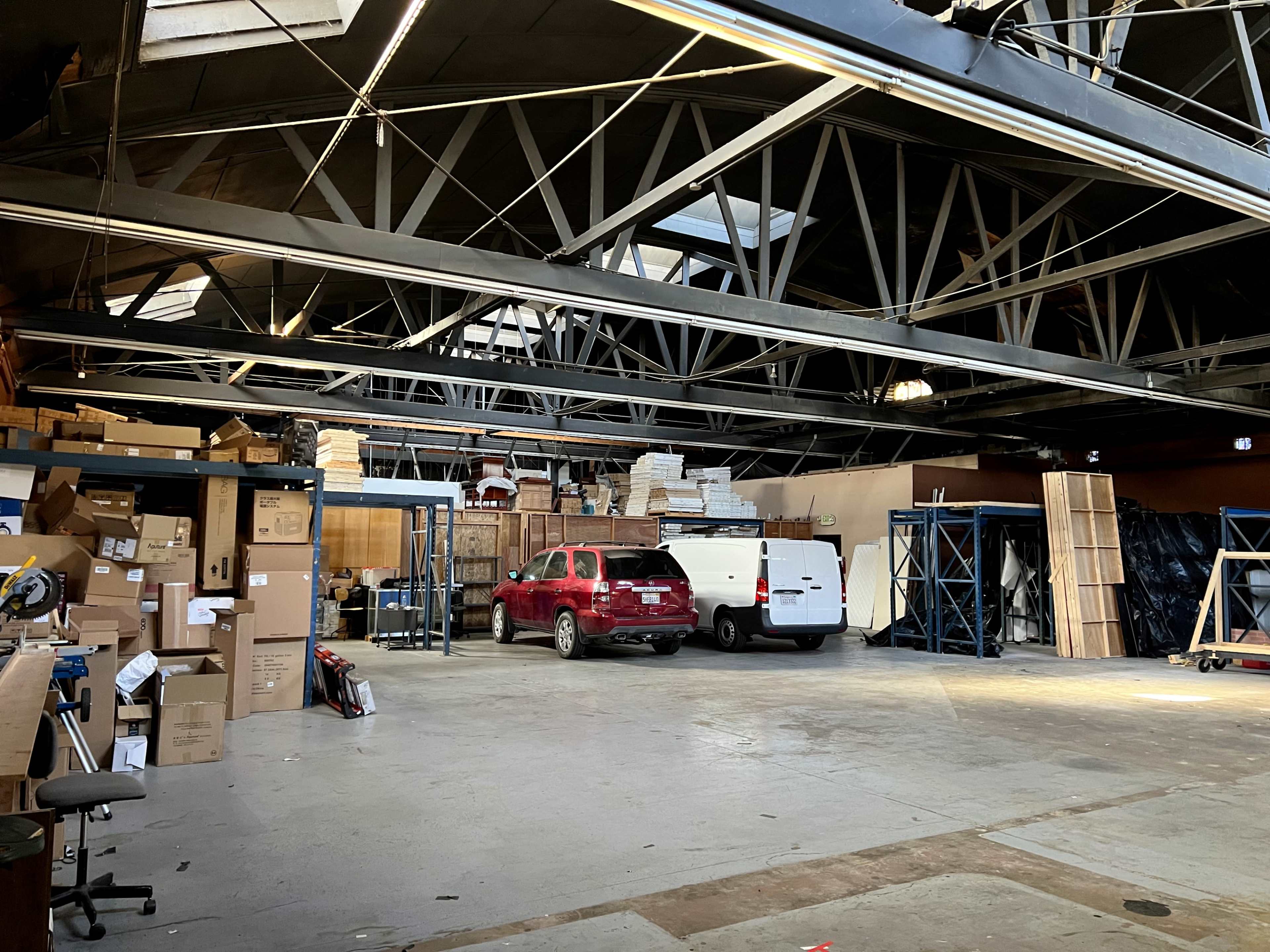 A large warehouse space containing stacked boxes, shelving units, and two vehicles parked inside.