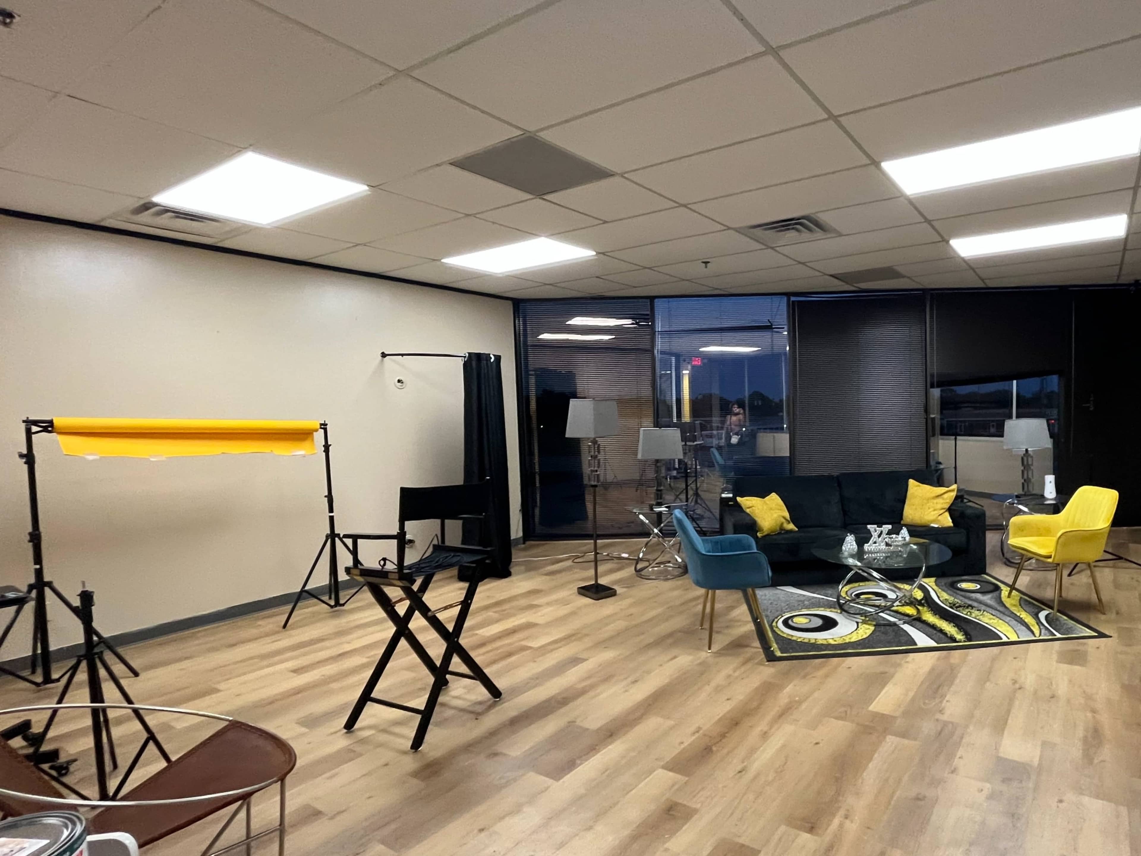 A modern studio space with a black sofa, yellow and blue chairs, lighting equipment, and a colorful rug on wooden flooring.