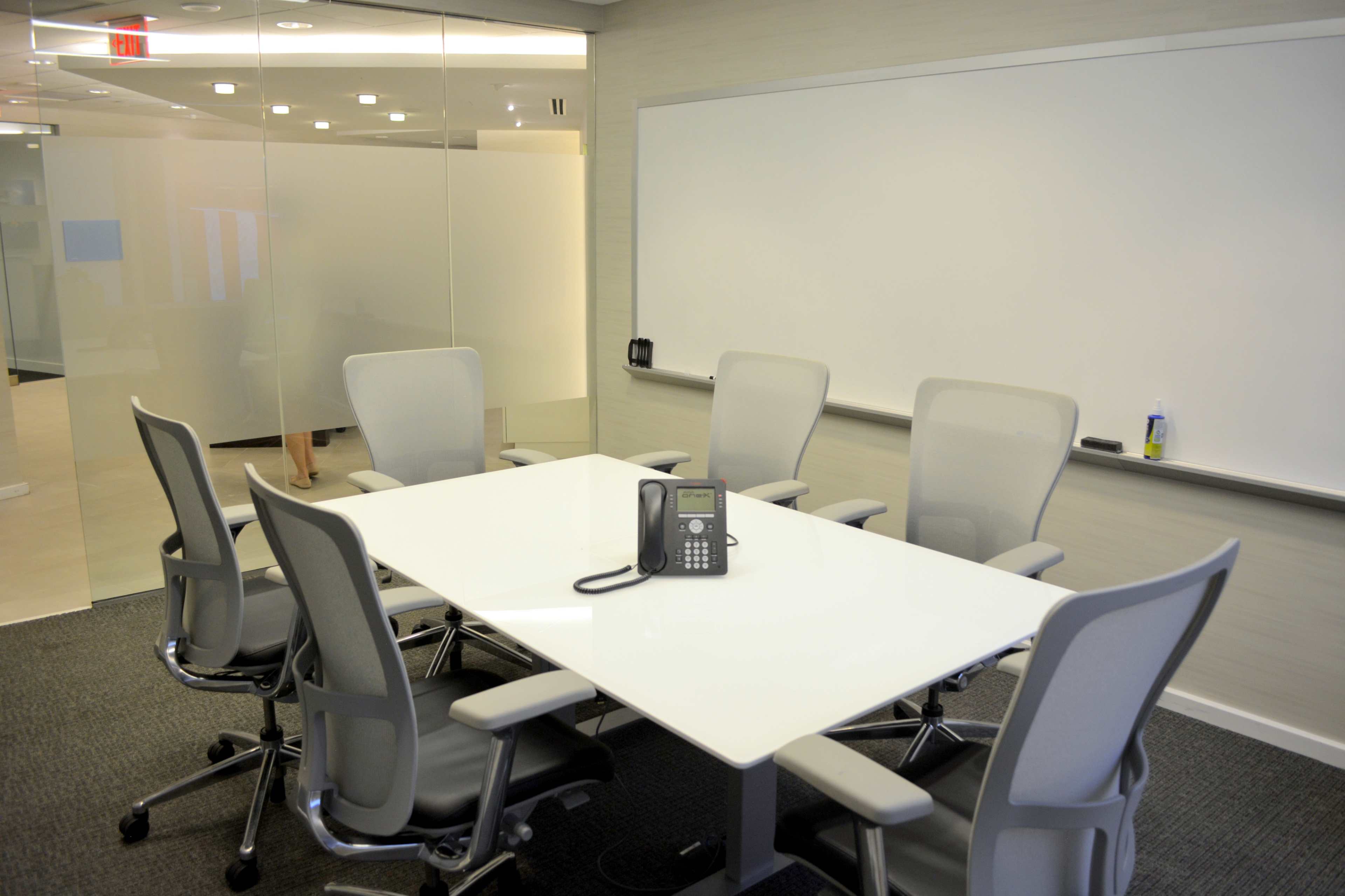 A conference room features a white table surrounded by five chairs, with a phone positioned in the center, and a whiteboard on one wall.