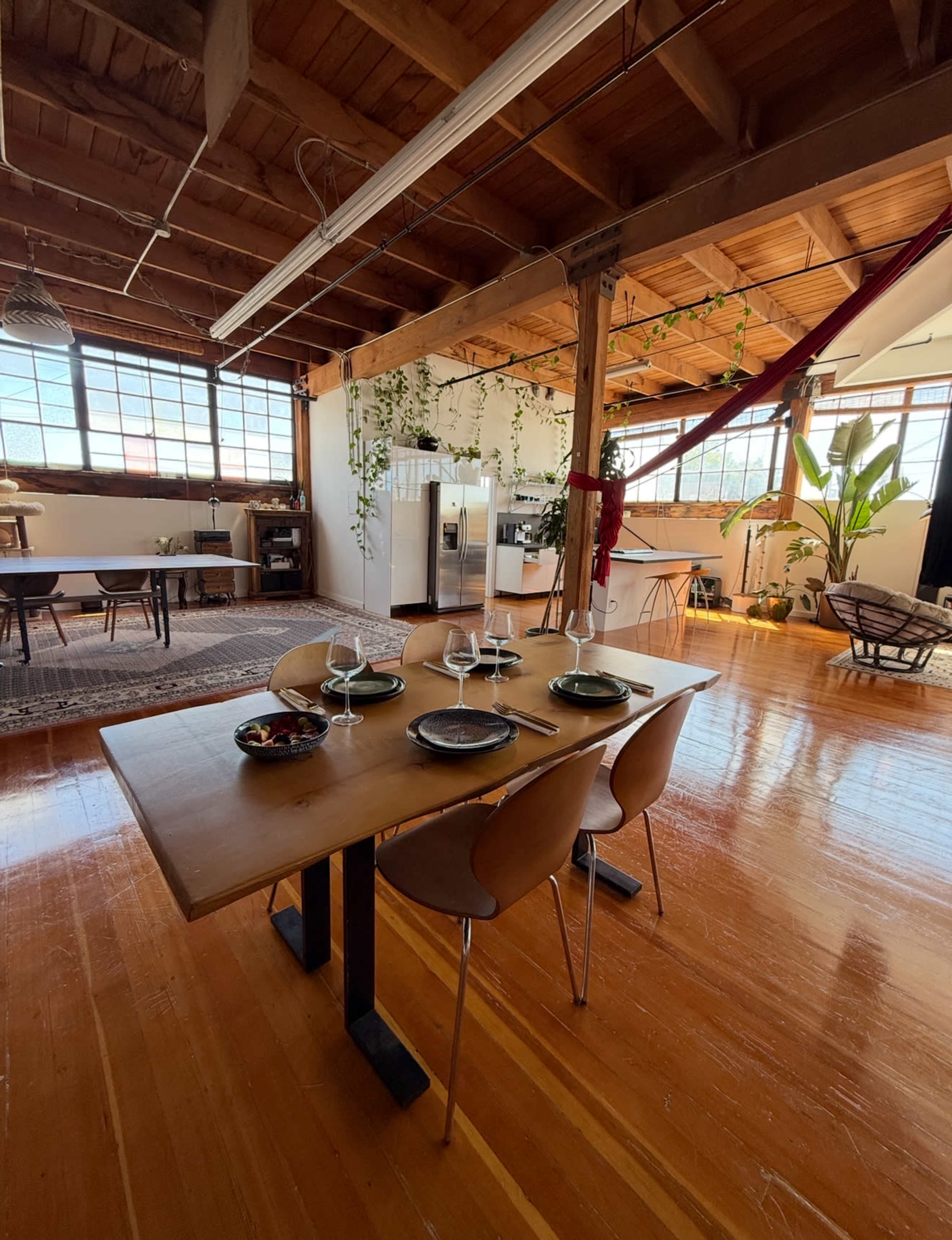The image shows a spacious dining area with a wooden table set for four, surrounded by large windows and plants in a well-lit open space.