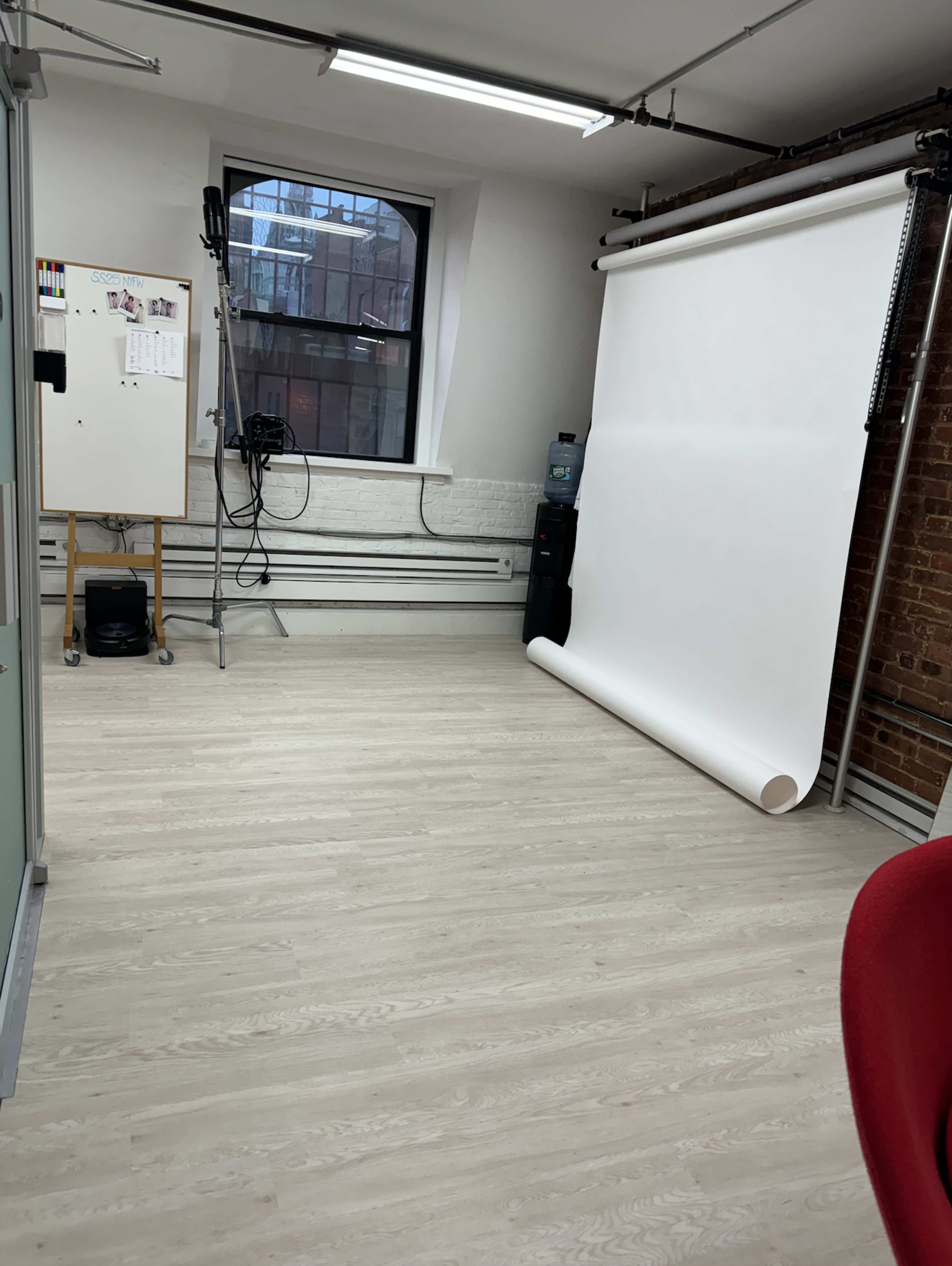 The image shows a brightly lit studio space with a white backdrop set against a brick wall, accompanied by a water cooler and a tripod stand on the floor.