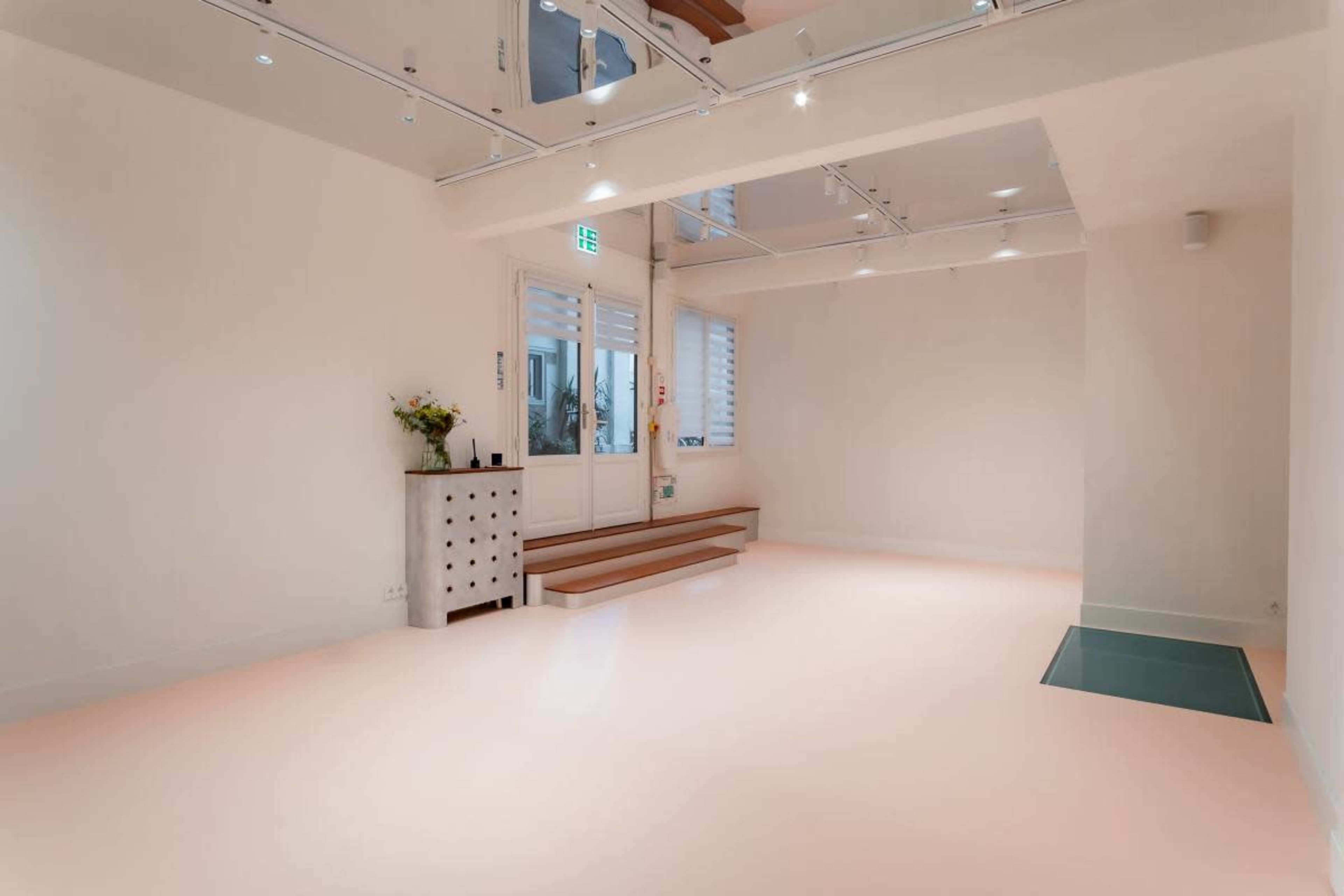 The image shows a minimalist, airy room with light-colored walls, wooden steps, and a small chest of drawers near a large window.