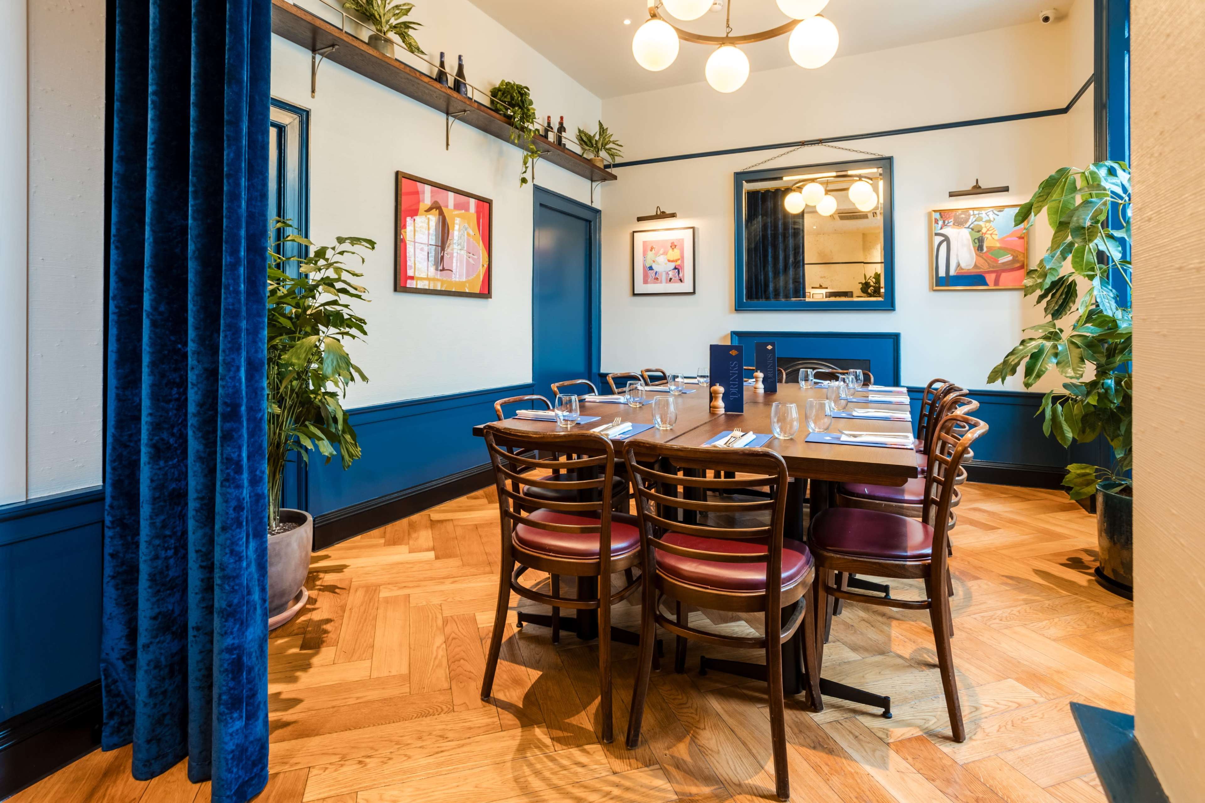 The image shows a dining area with a wooden table set for a meal, surrounded by chairs, decorative plants, and framed artwork on the walls.