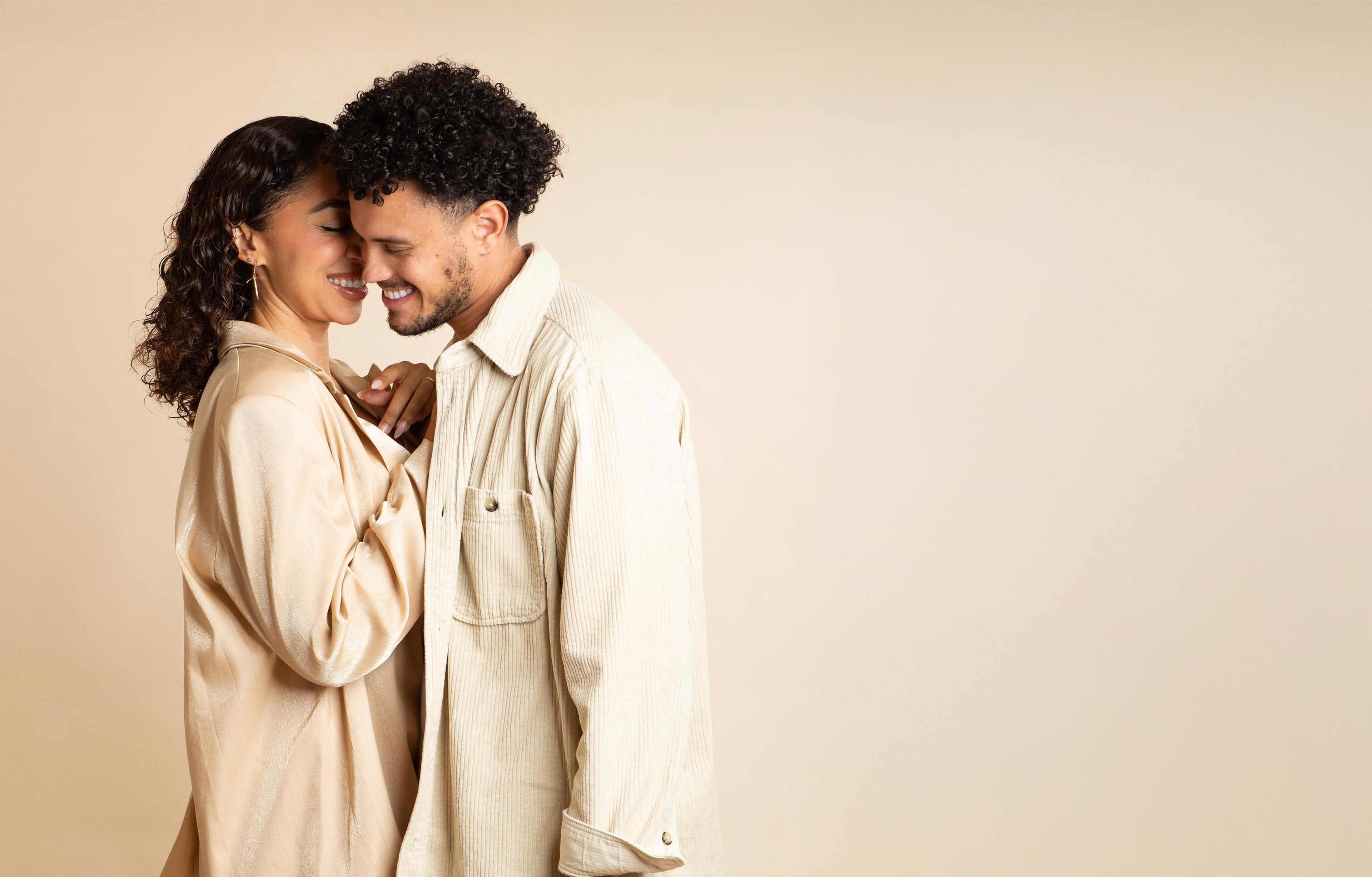A man and woman stand close together, smiling at each other against a plain, light-colored background.