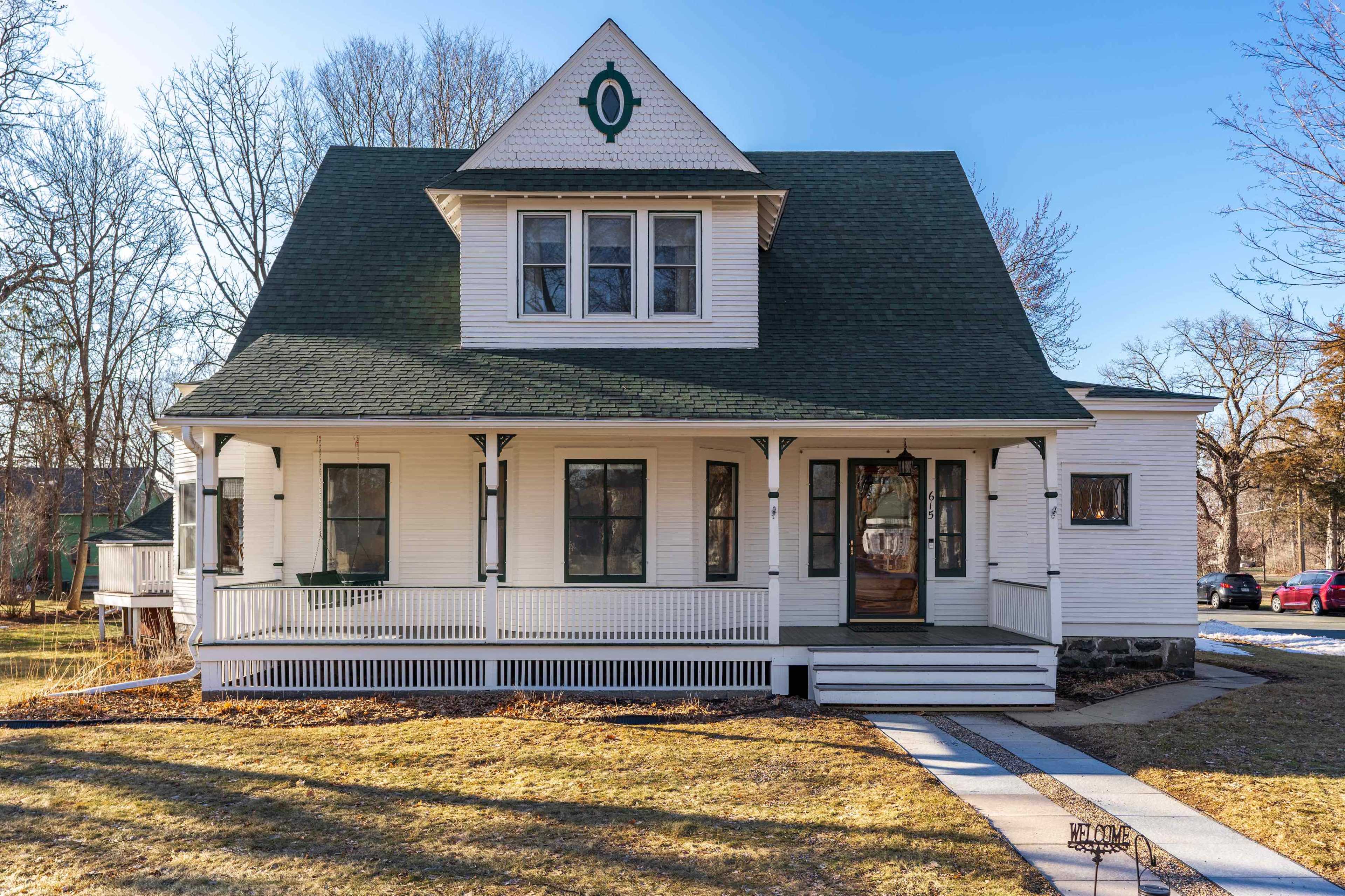 A two-story white house with a green roof and decorative elements stands on a grassy lawn, featuring a front porch and multiple windows.