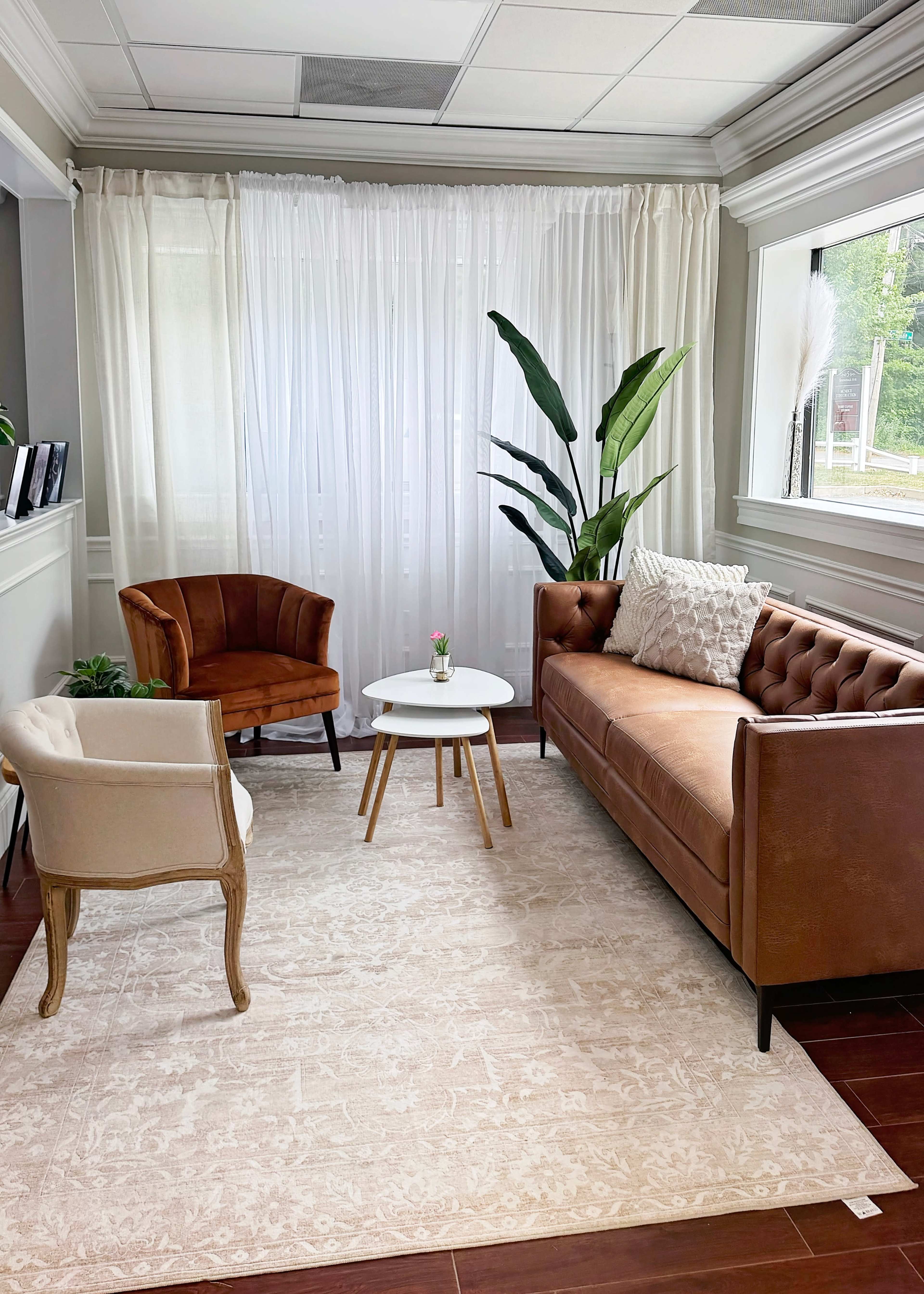 The image shows a seating area with a brown tufted sofa, two accent chairs, a small white coffee table, and a potted plant, all arranged on a light-colored rug near a large window with sheer curtains.