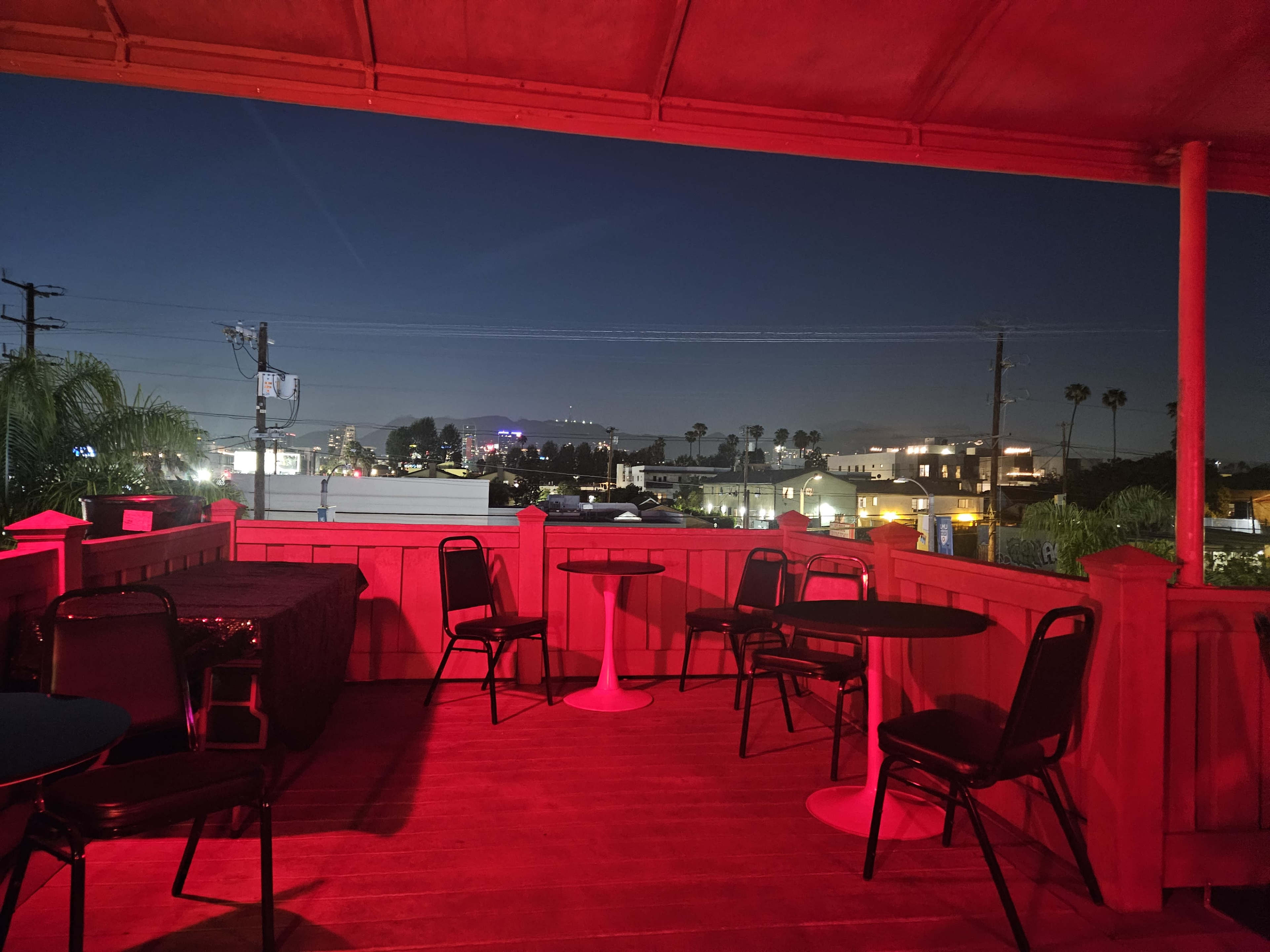 The image shows a rooftop space with red lighting, empty tables and chairs, and a view of a distant cityscape at twilight.
