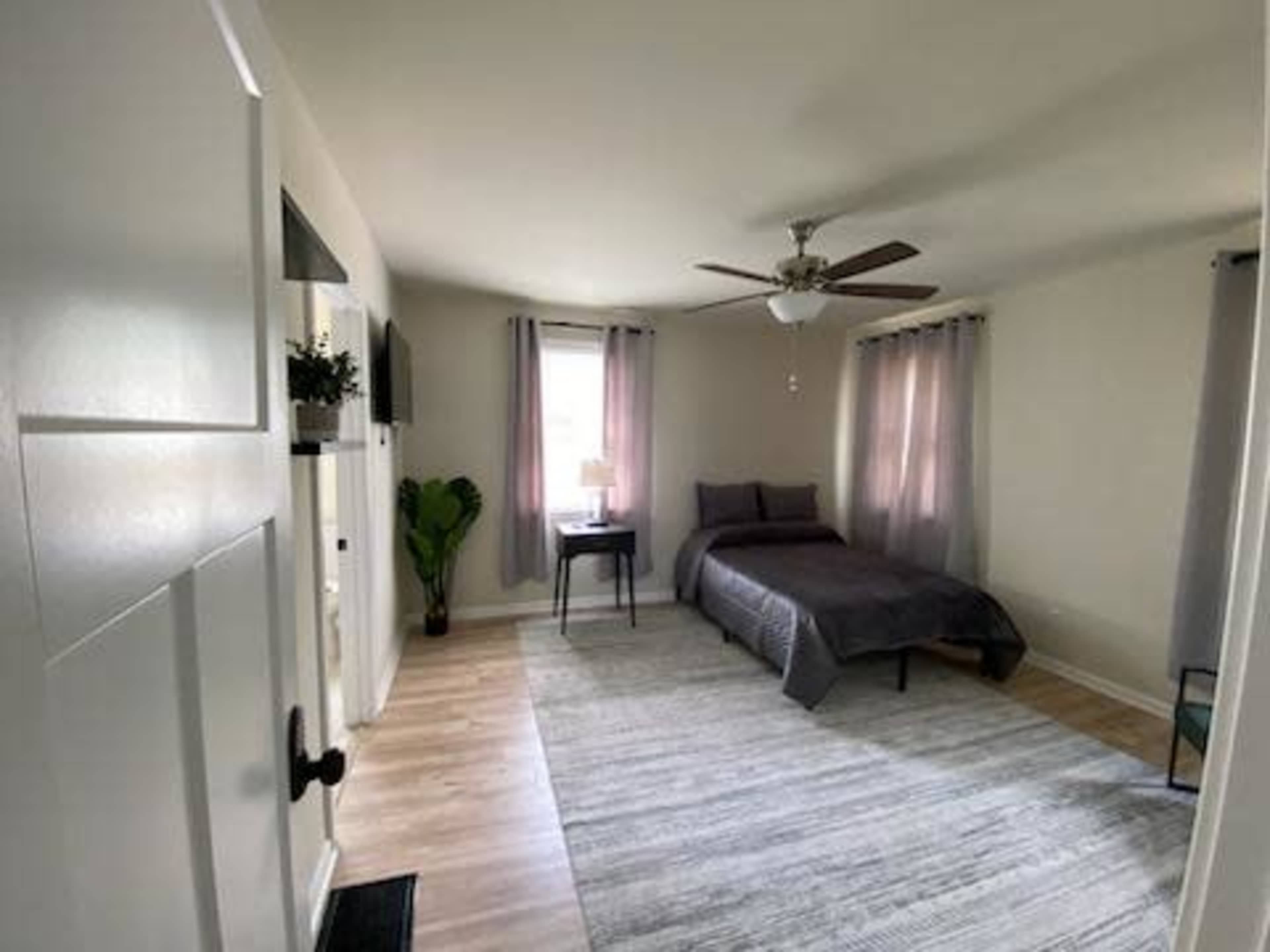 A simple bedroom with a bed, a small table, a ceiling fan, and large windows with curtains.