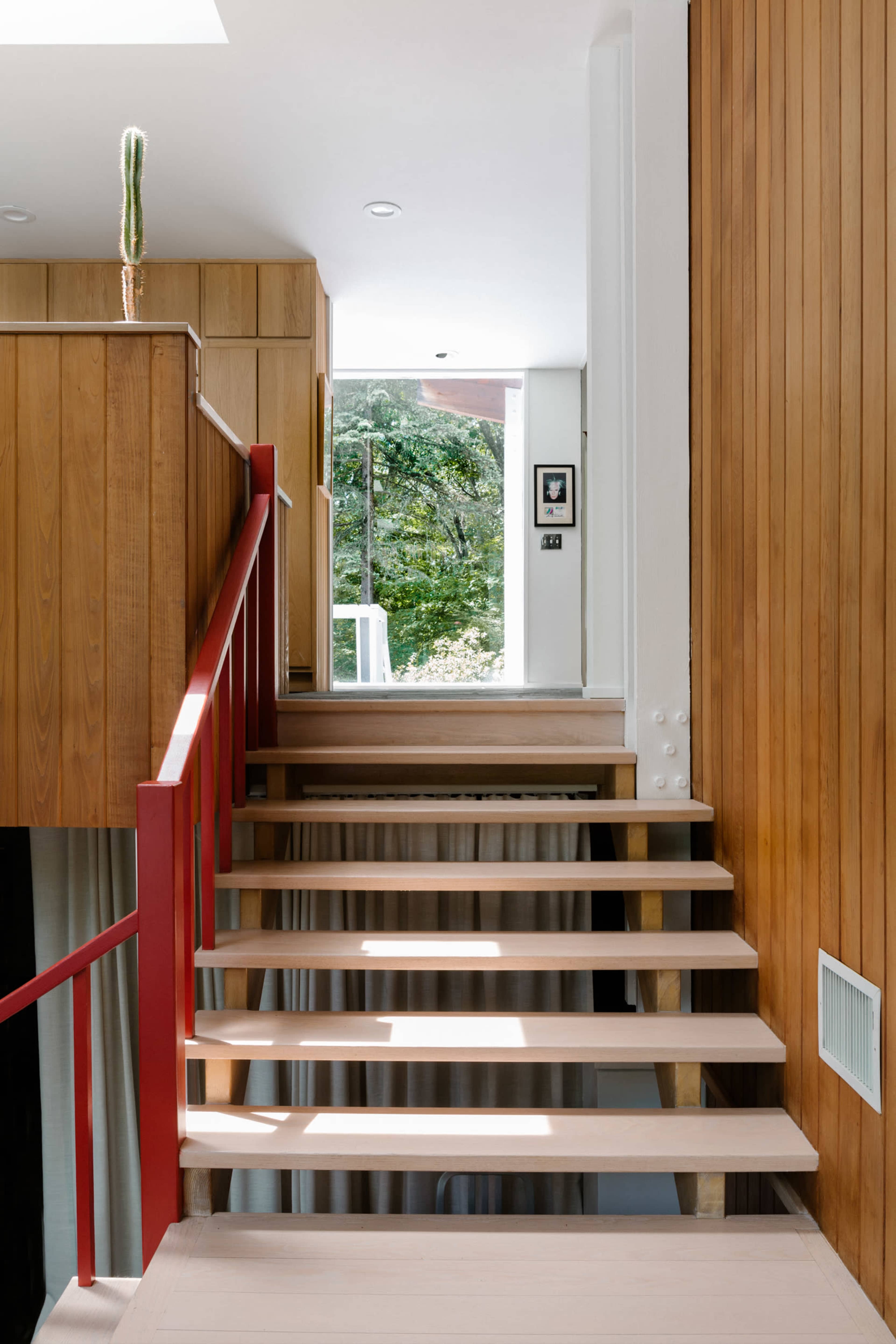 A staircase leading to a bright upper level, with wooden and red railings, surrounded by natural light and greenery visible through a window.