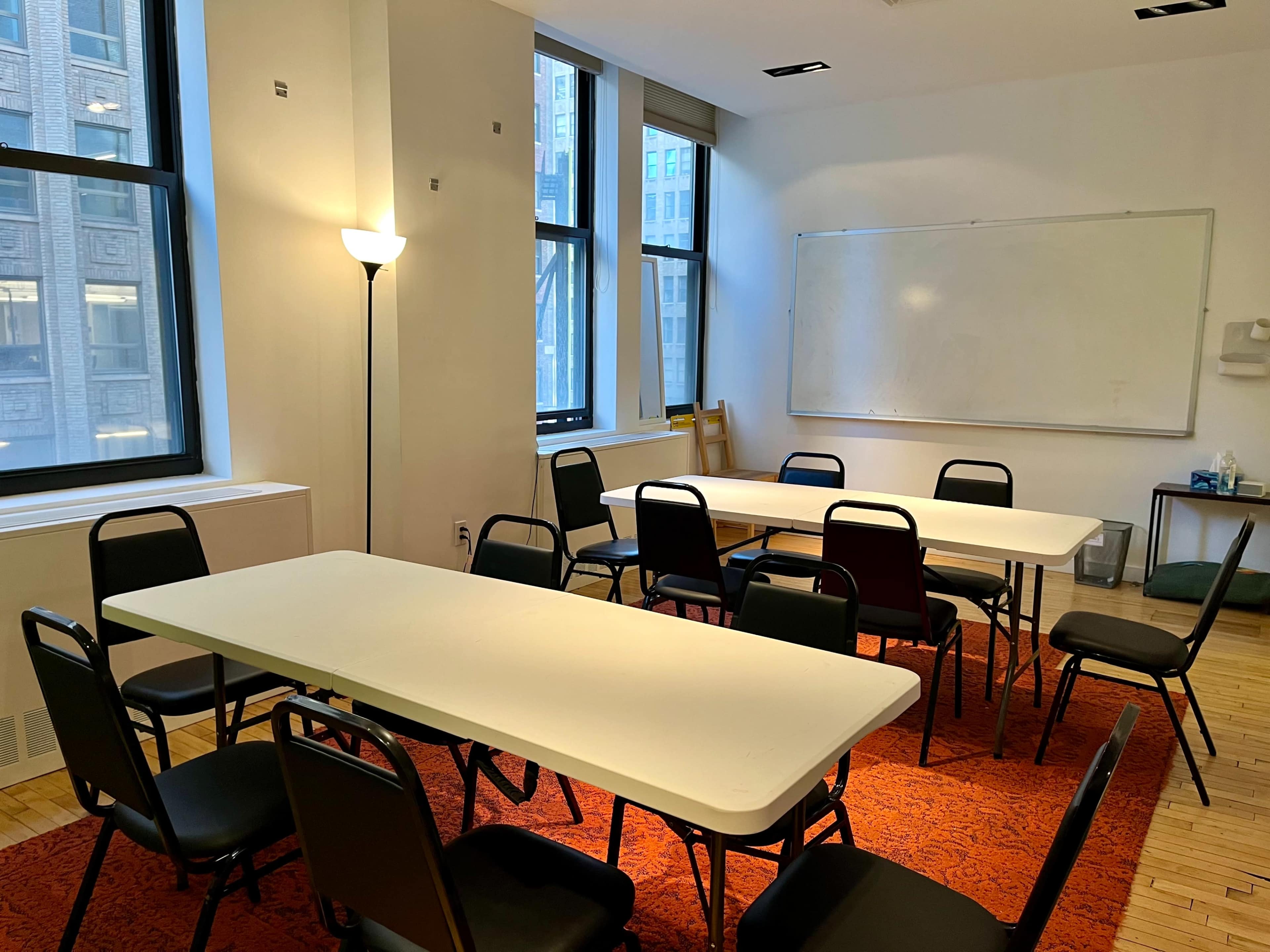 A meeting room features multiple white tables arranged in a circular layout surrounded by black chairs, with large windows allowing light to enter.