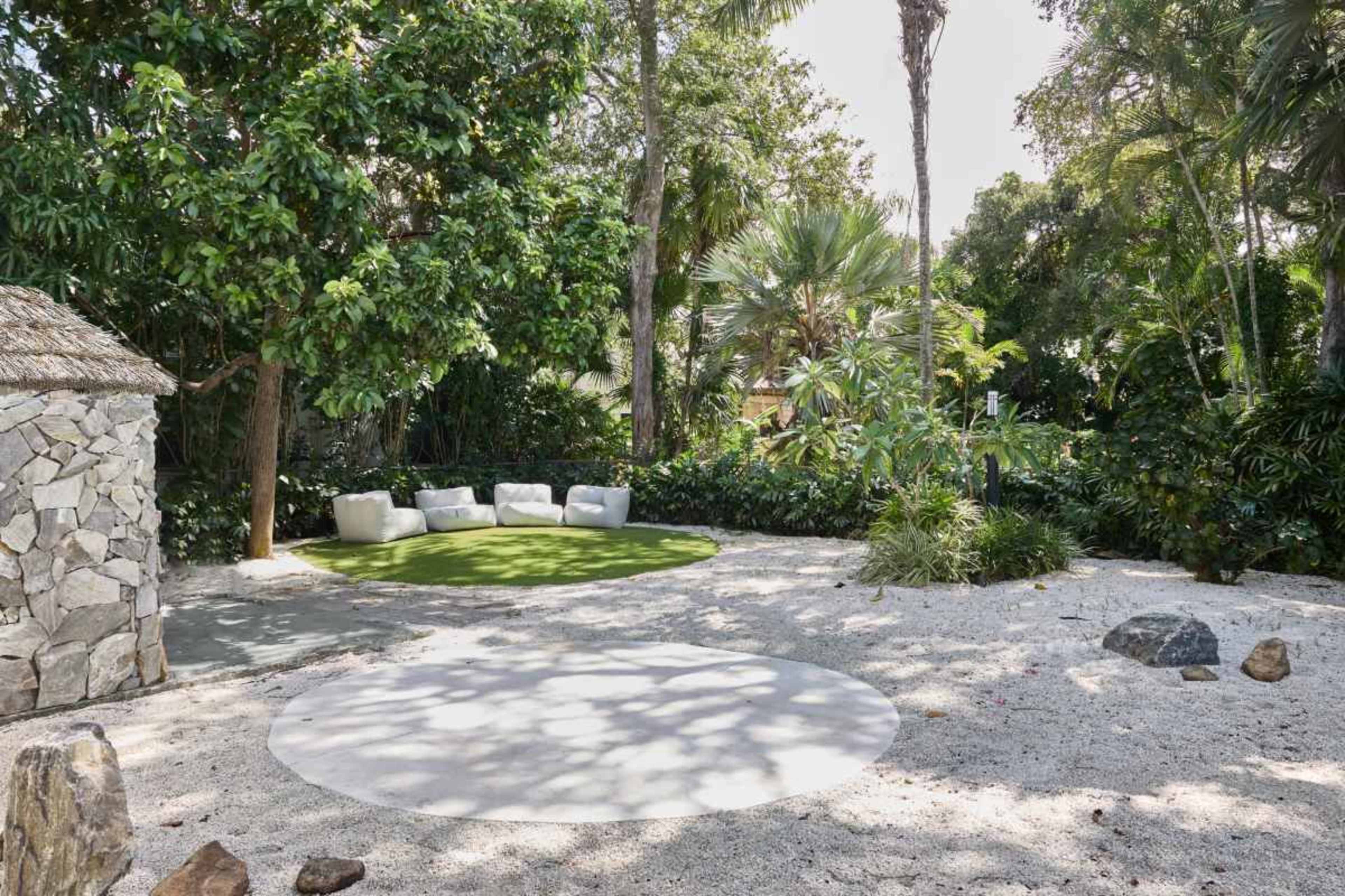 The image shows a landscaped outdoor area featuring a circular stone patio, a grassy section with white couches, and lush greenery surrounding a small stone structure.