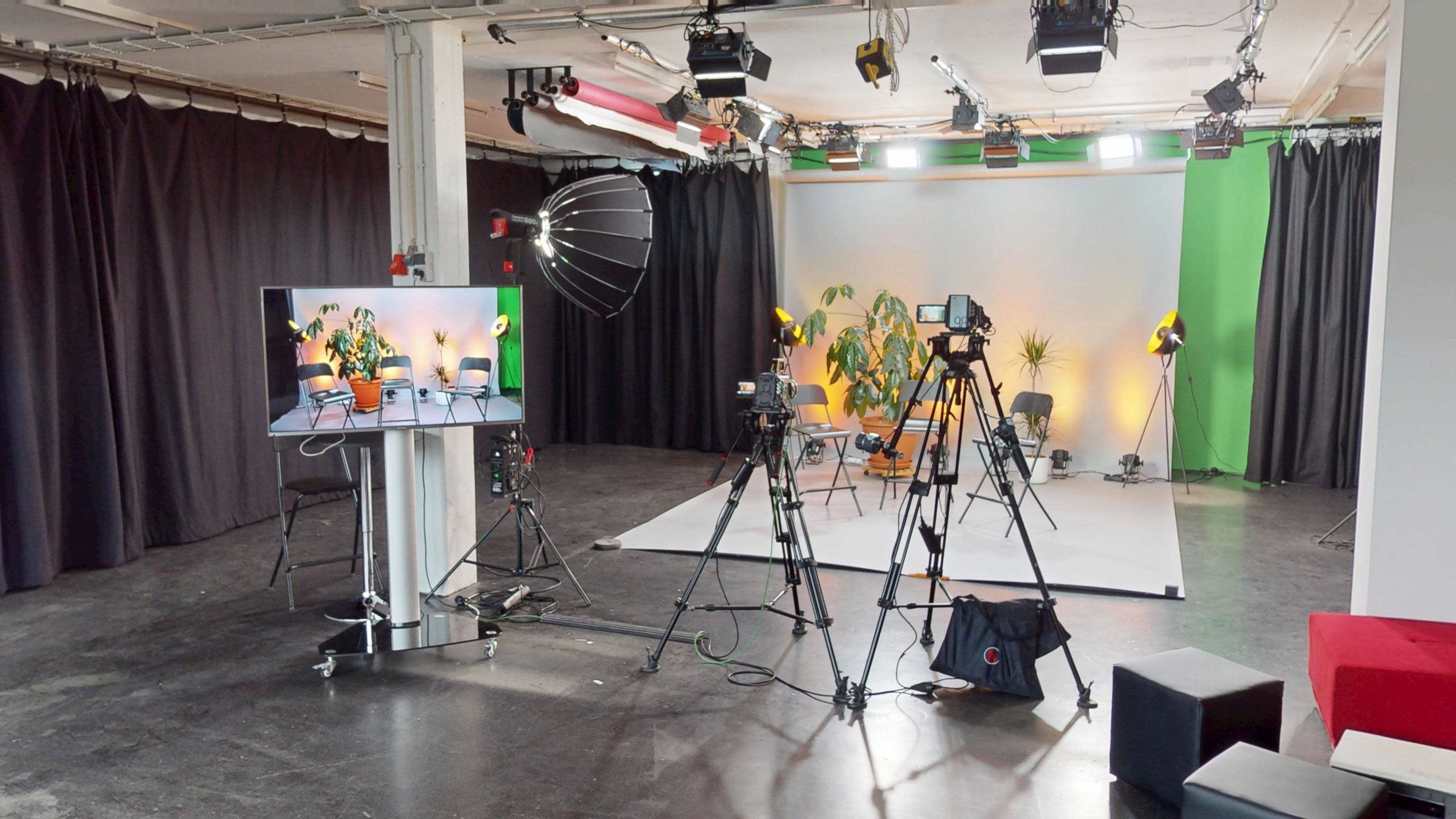 The image shows a video production studio equipped with multiple cameras, lighting setups, and a large screen displaying a video call or interview setup.