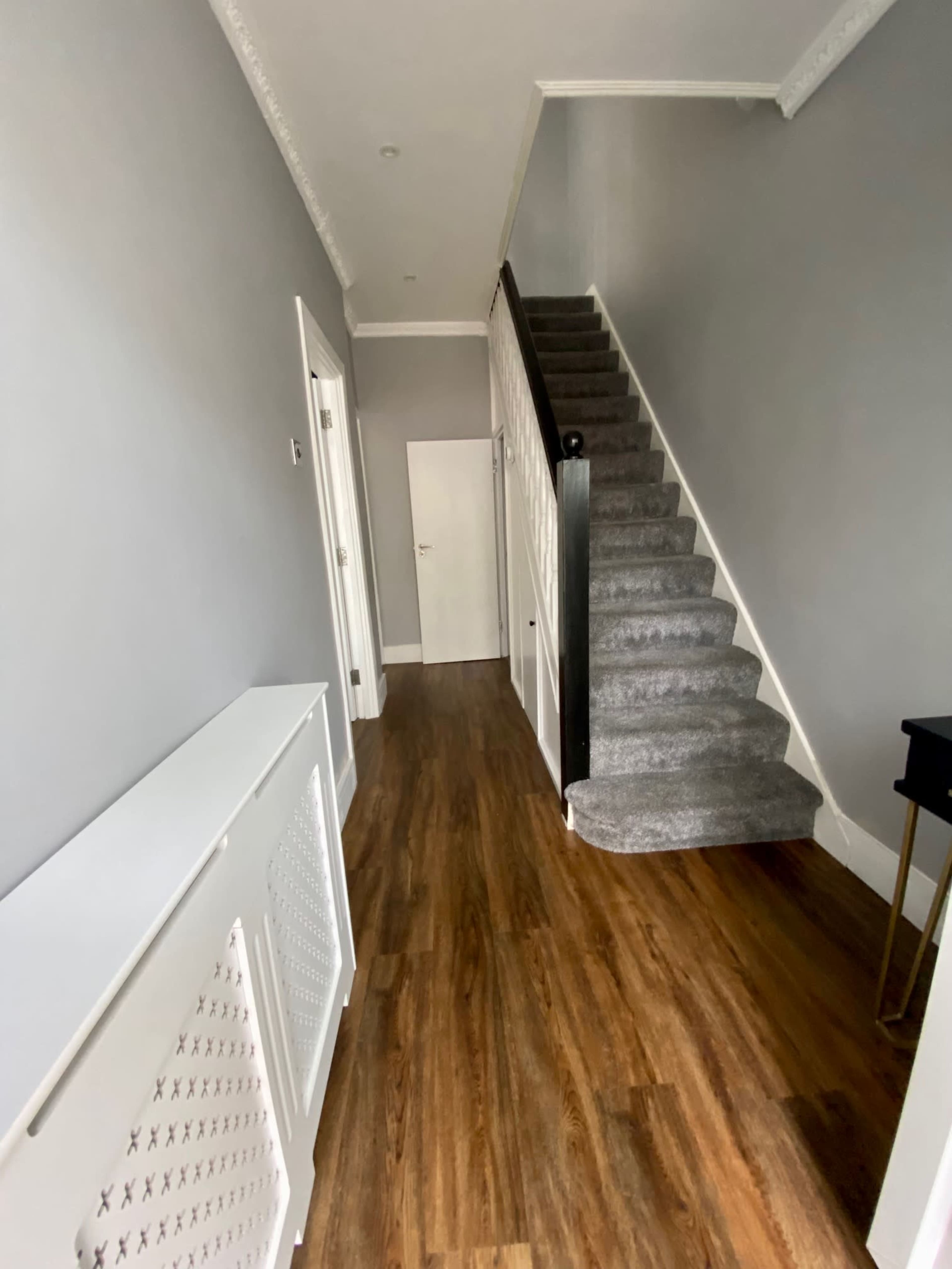 A long hallway with a staircase leading up, gray walls, wooden flooring, and a white radiator cover along one side.