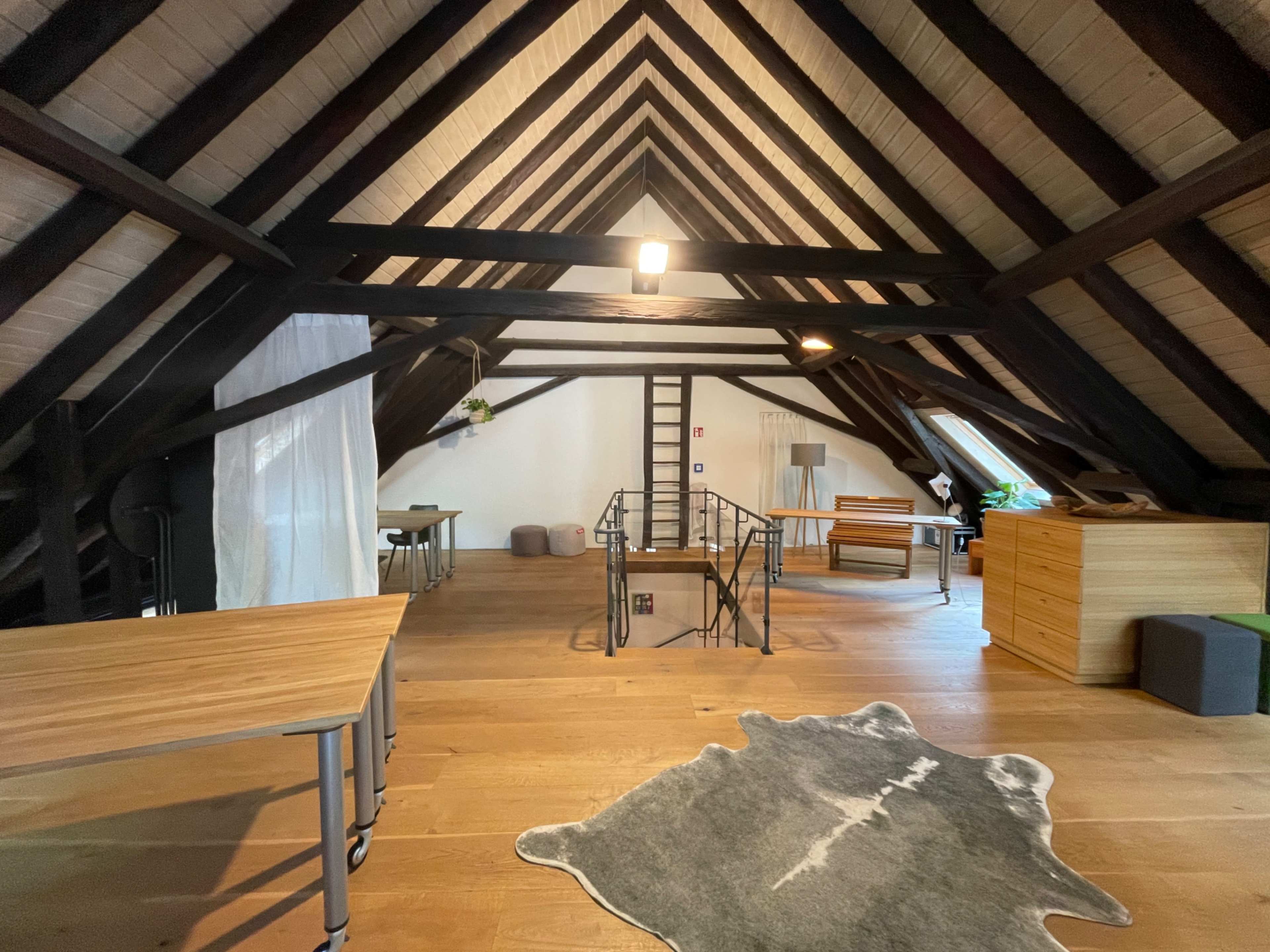 The image shows a spacious attic room with exposed wooden beams, wooden flooring, and various furniture pieces, including tables and a couch.