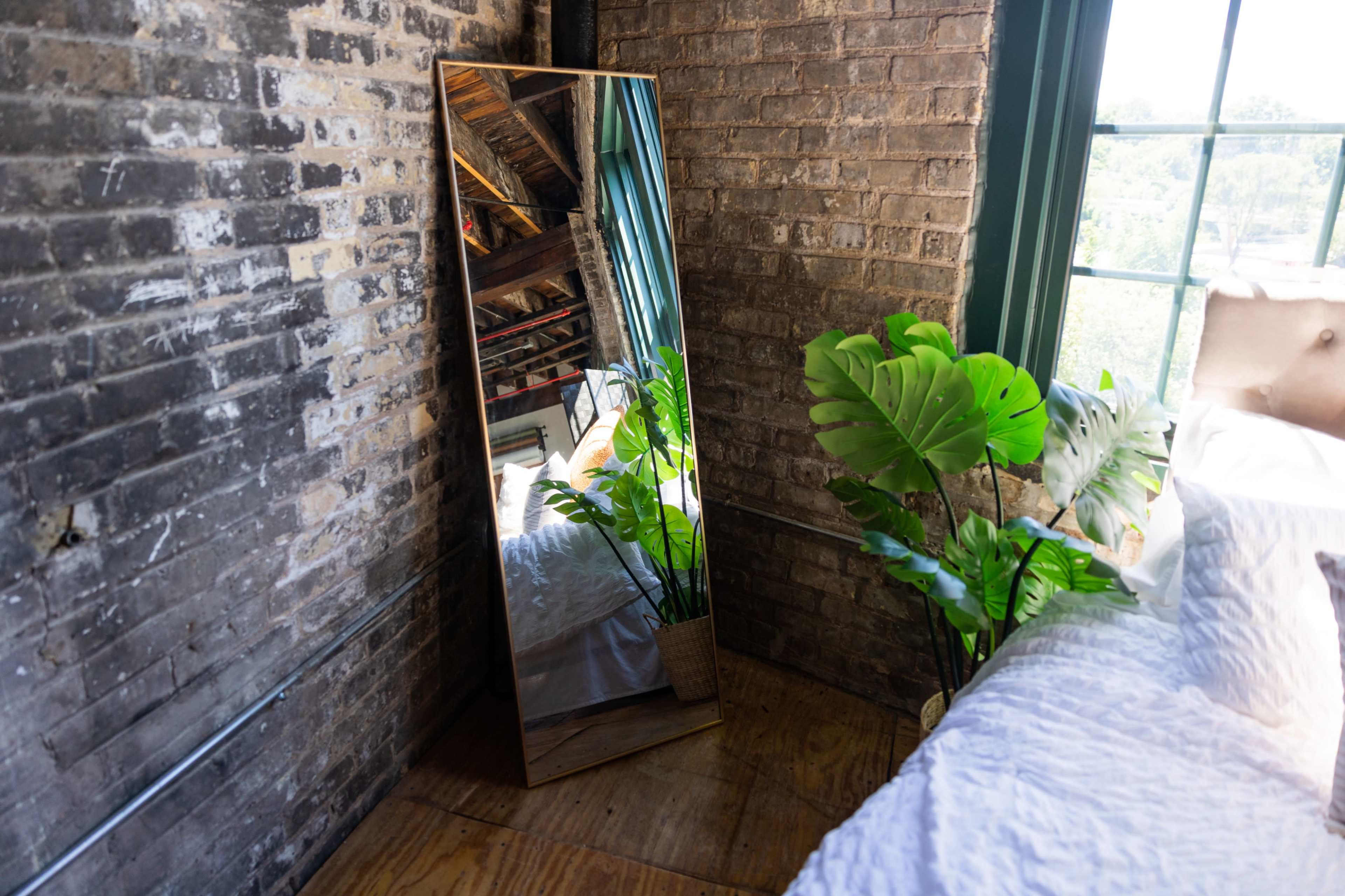 A tall mirror leans against a brick wall beside a potted plant in a room with wooden flooring.