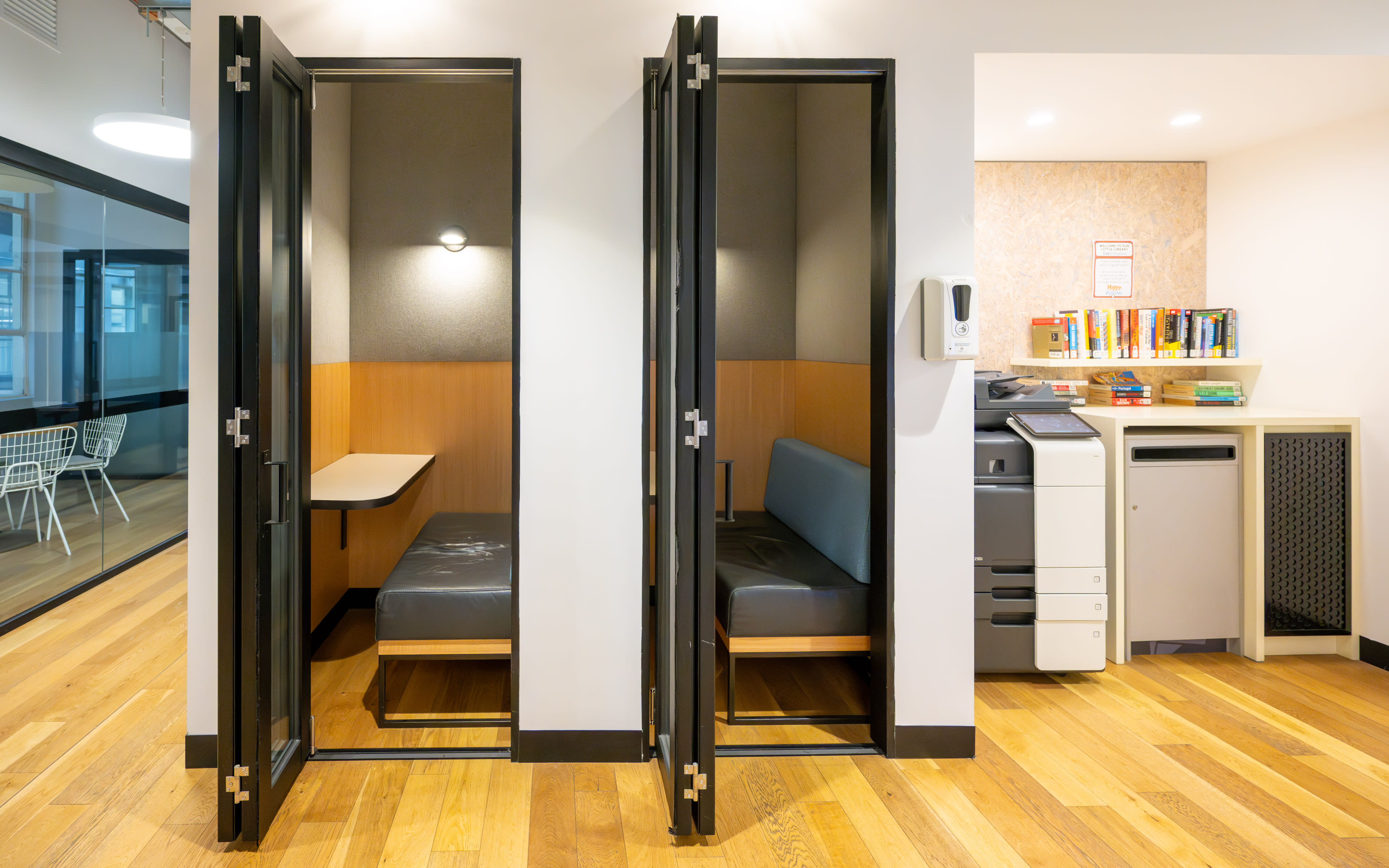 Two open soundproof booths with seating against the wall, adjacent to a work area featuring a printer and bookshelves.