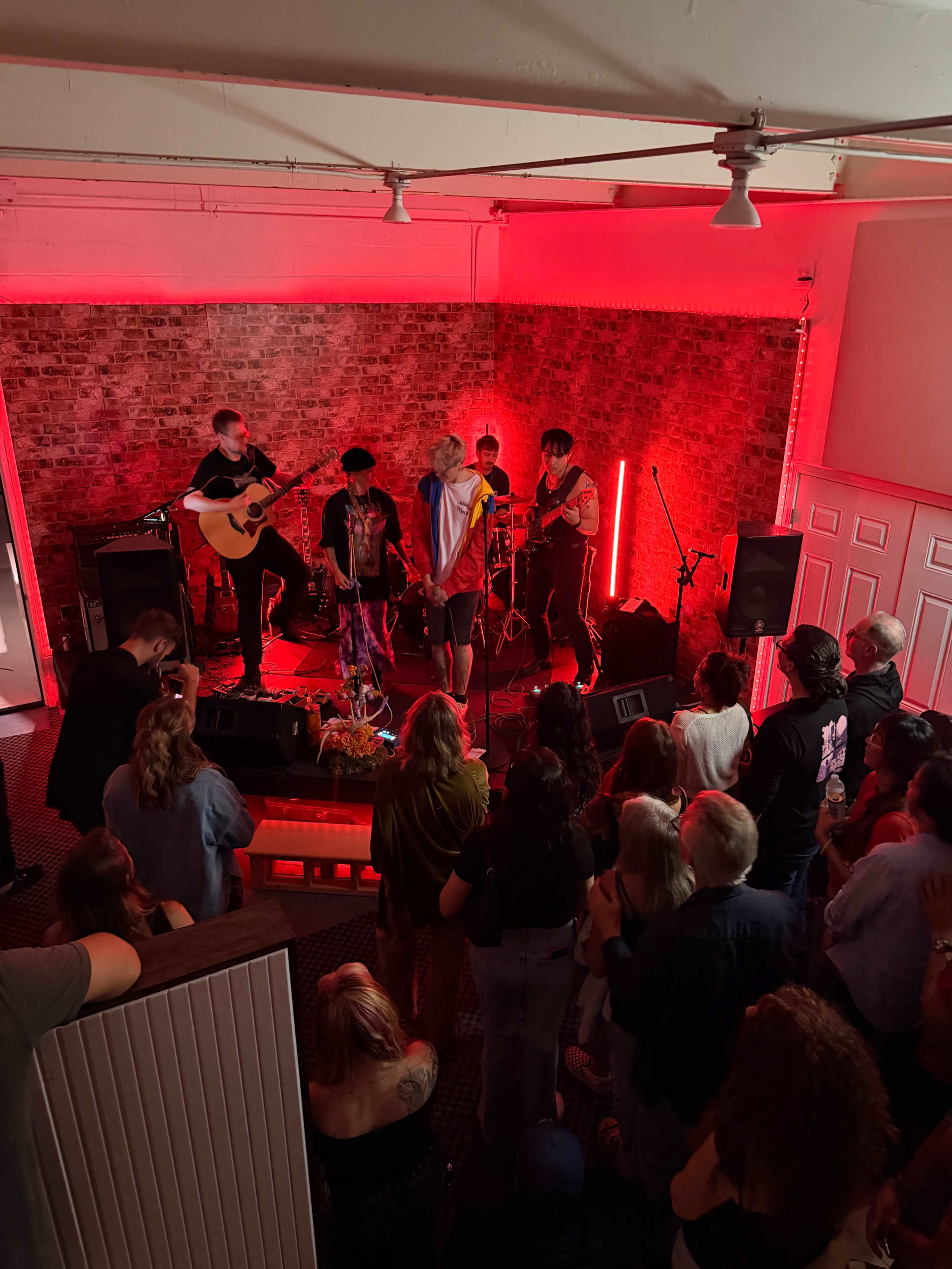 A band performs on stage in a dimly lit venue with red lighting, while an audience watches from the floor.