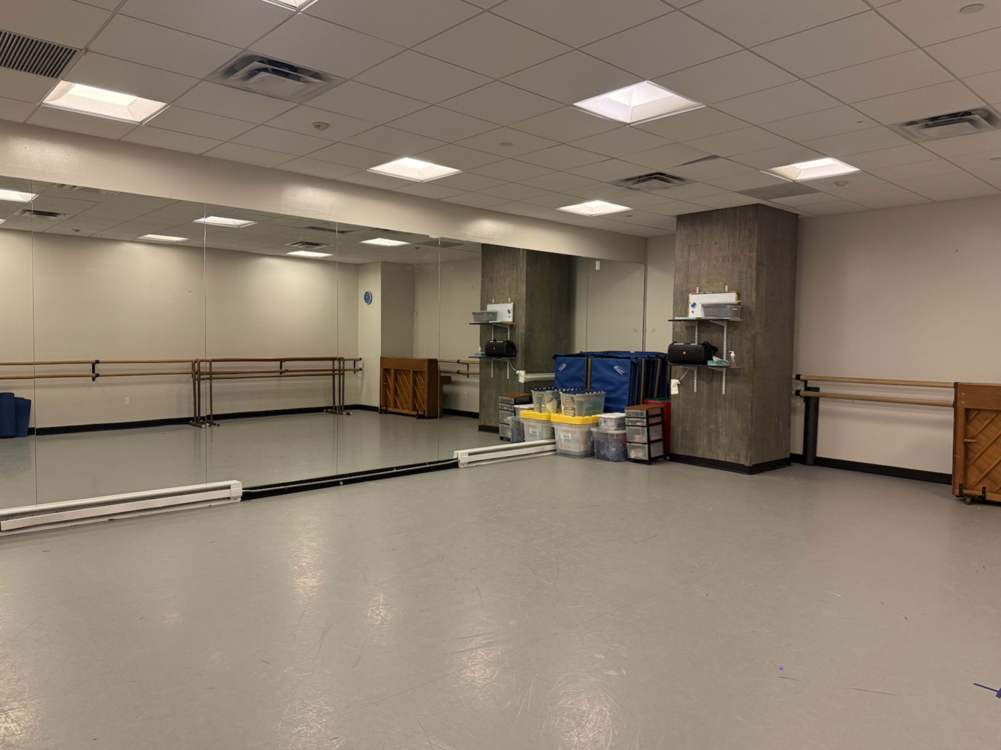 The image shows a dance studio with mirrored walls, a ballet barre, and storage bins arranged in the corner.