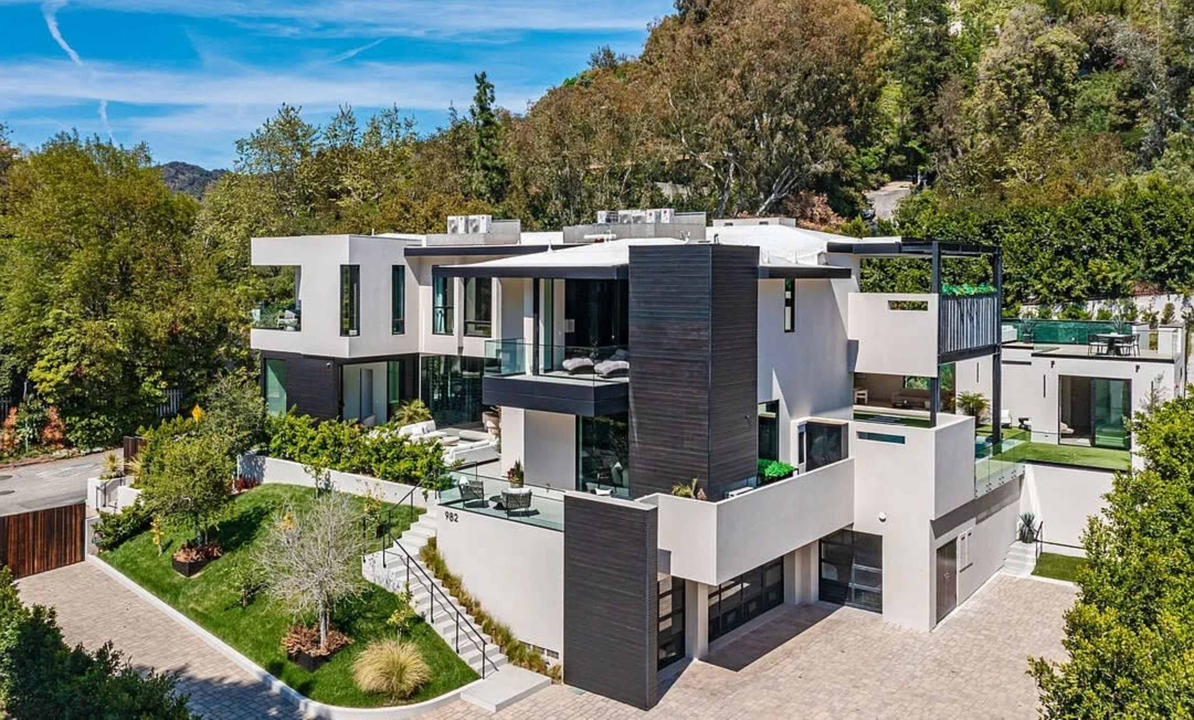The image shows a modern, multi-story house with large windows and a flat roof, surrounded by greenery and a paved driveway.