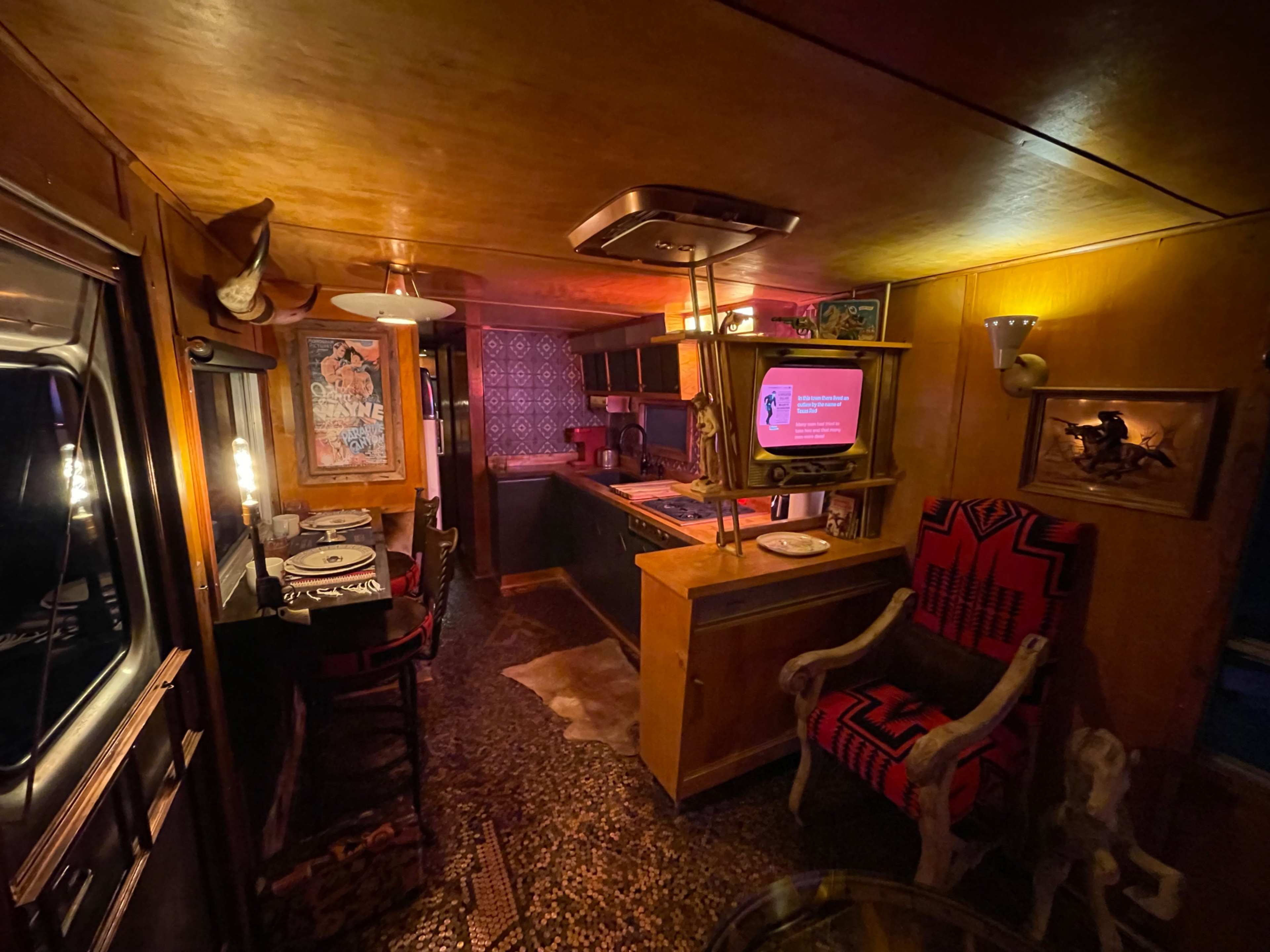 The image shows an interior of a vintage-styled kitchen and dining area featuring wooden paneling, patterned wallpaper, and eclectic decor.