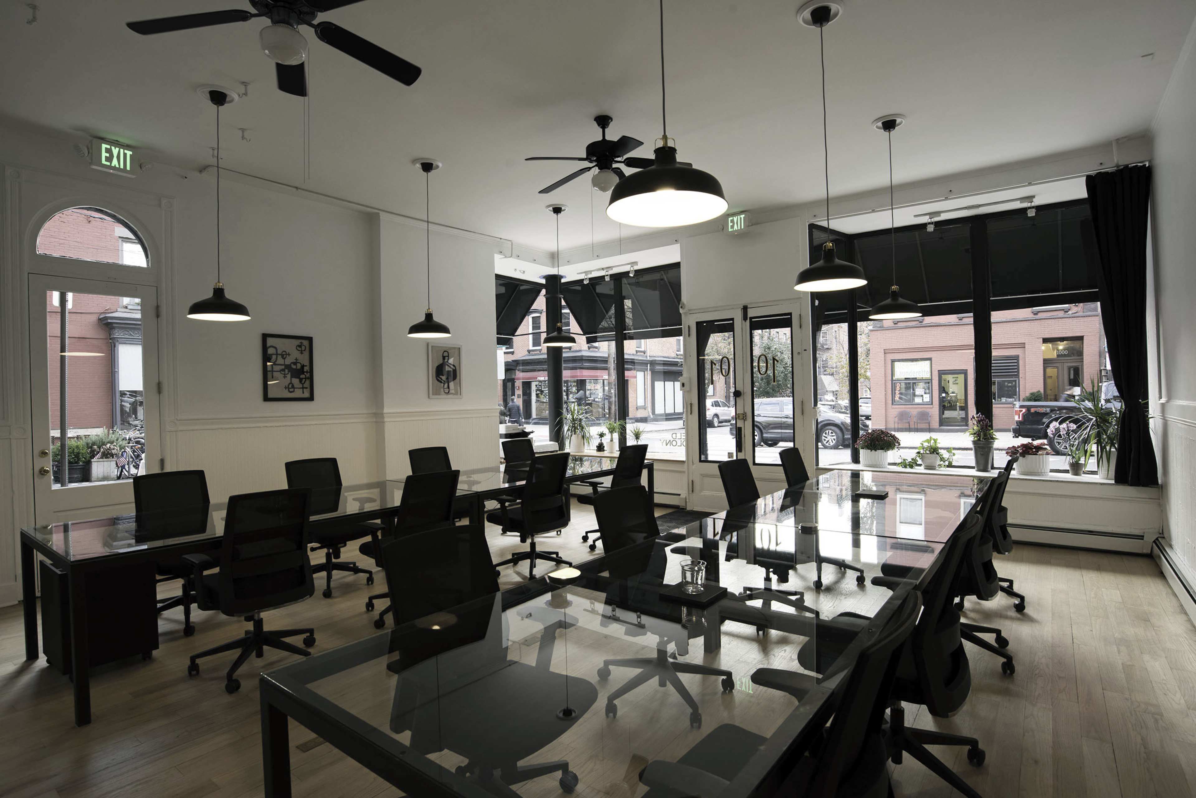 A modern office space with several glass-top tables and black chairs, illuminated by overhead lights and large windows facing an exterior street.