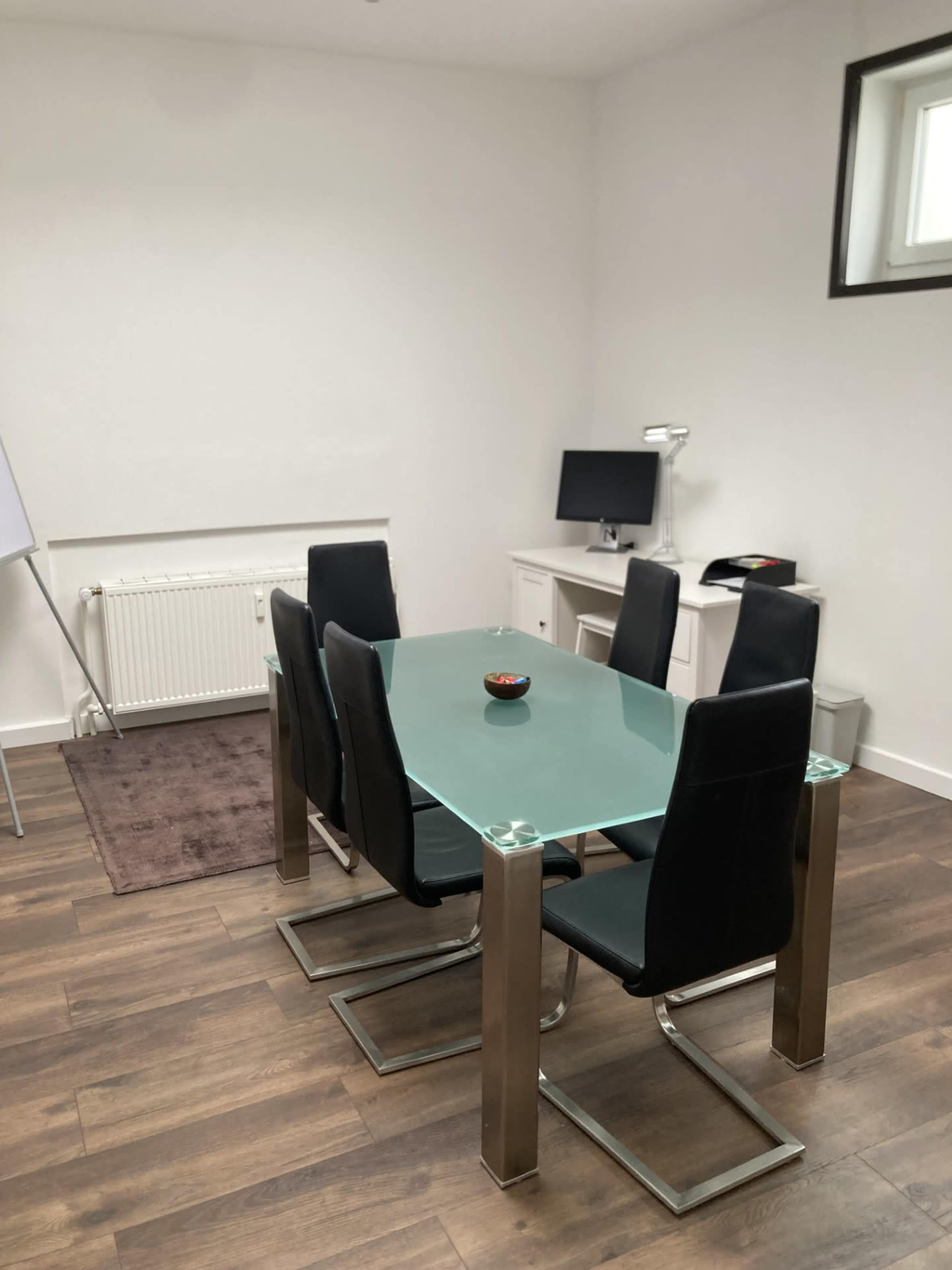 The image shows a minimalist meeting room with a glass dining table surrounded by black chairs, a monitor on a desk, and a radiator.