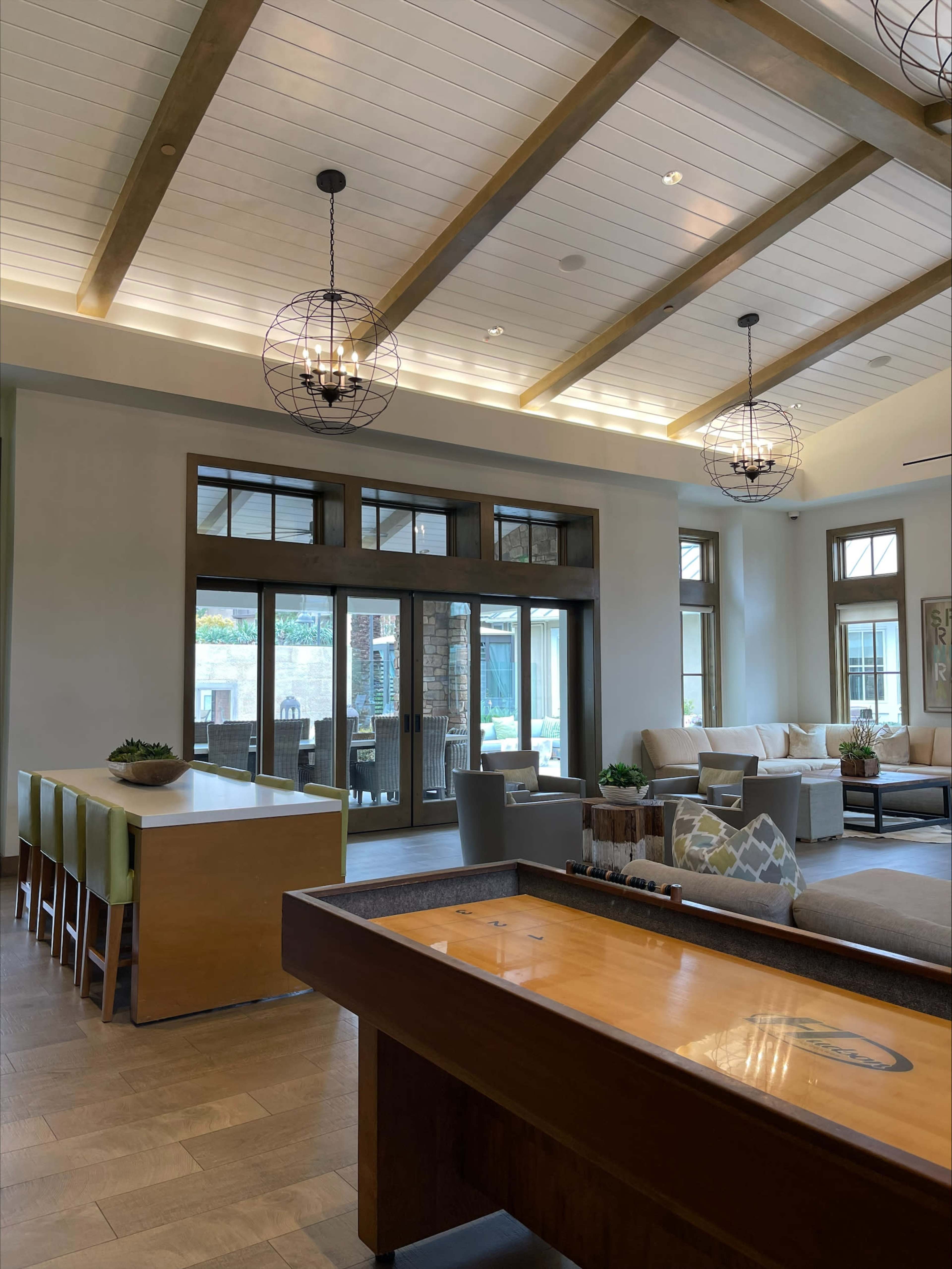 A modern lounge area featuring a shuffleboard table, seating arrangements, and large windows allowing natural light.