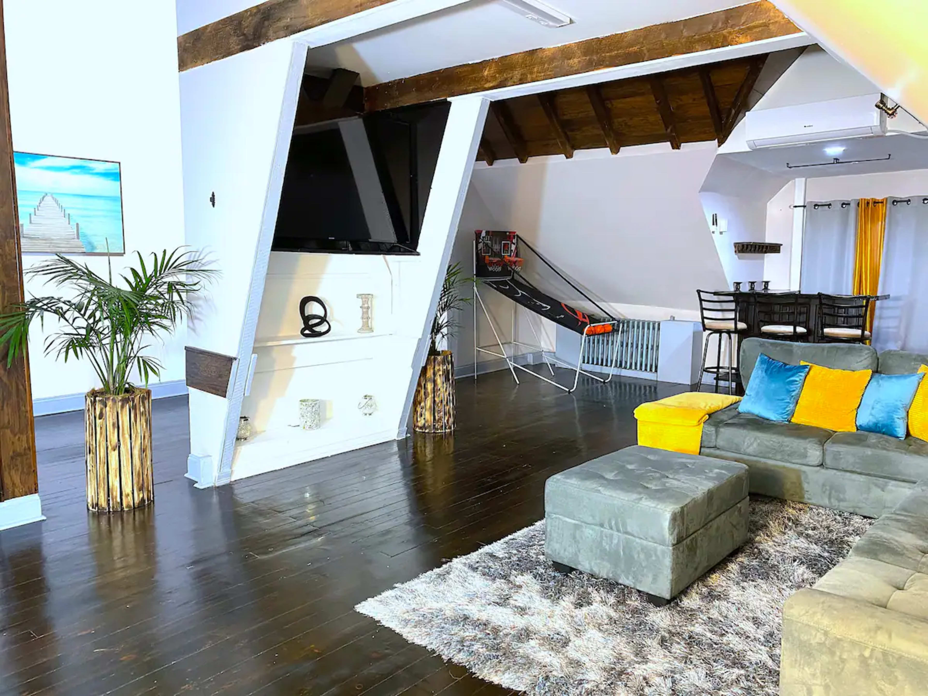 A modern living space with a gray sectional couch, a flat-screen TV mounted on a slanted wall, and a dining area adjacent to it.