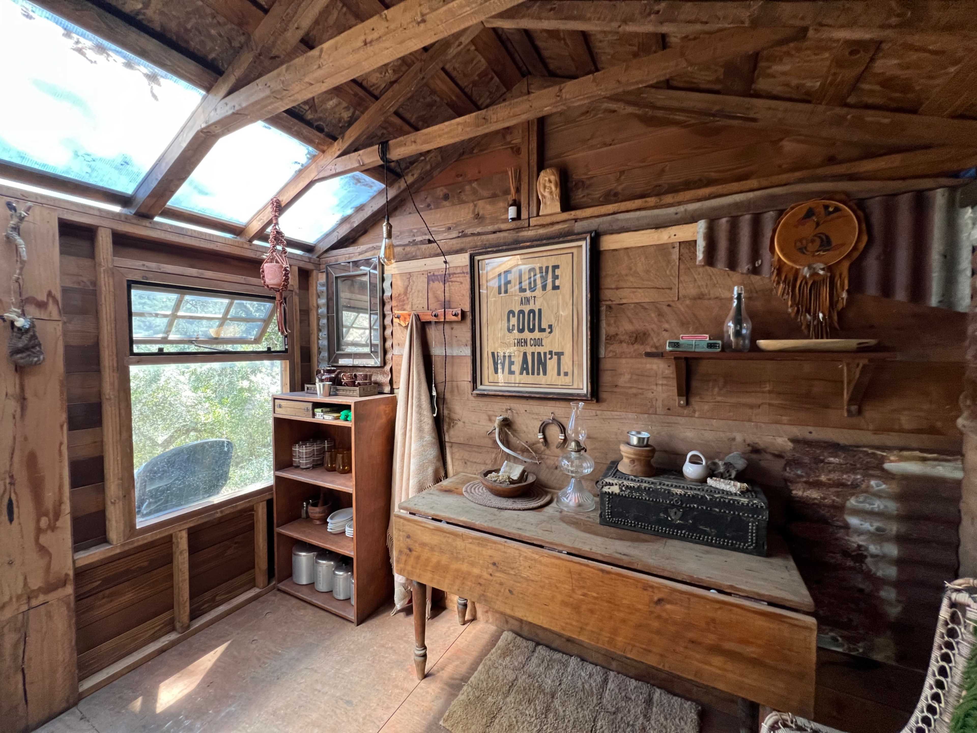 The interior of a rustic cabin features wooden walls, a table with various objects, and a large window allowing natural light to enter.